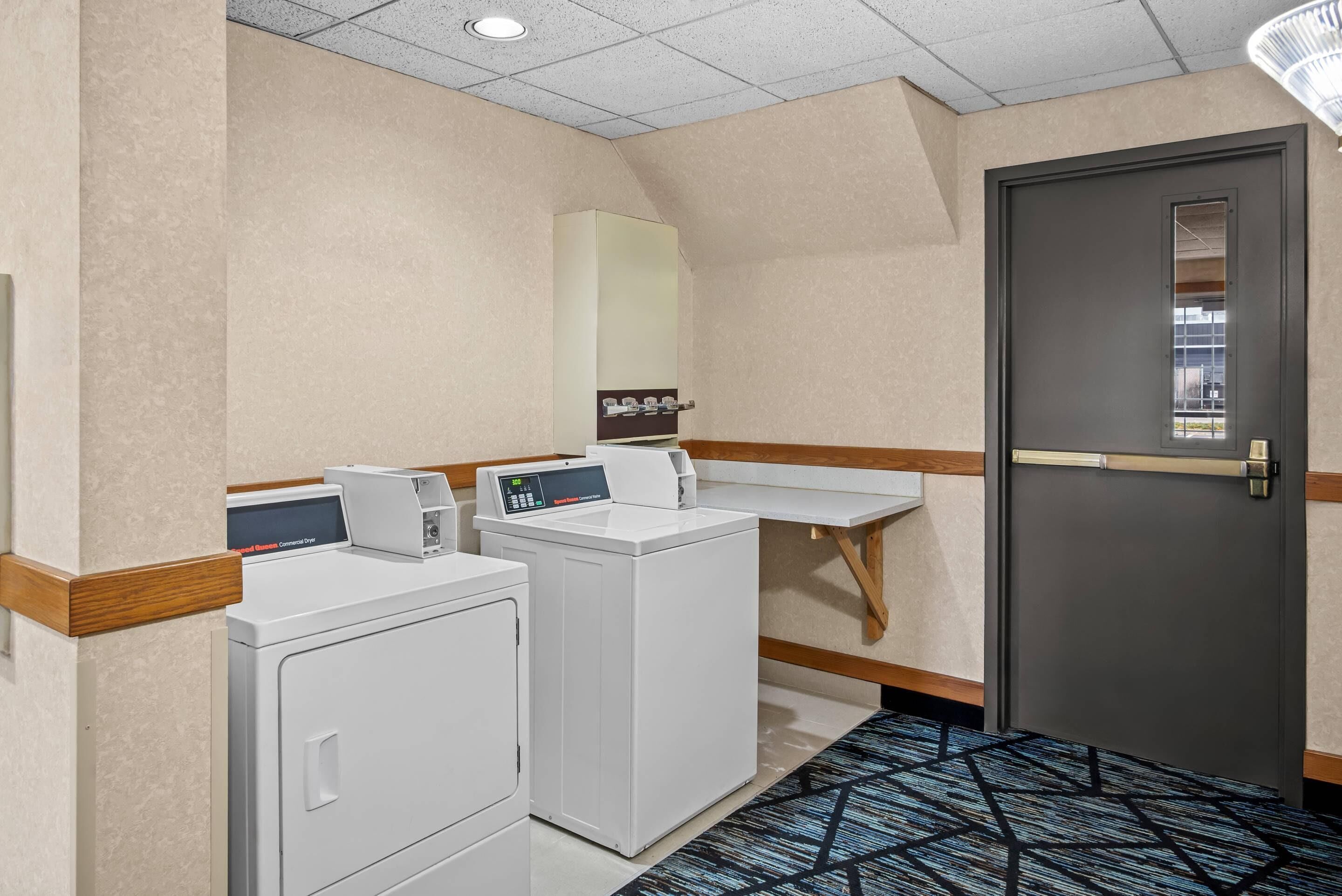 A hotel laundry room with two front-loading washers and dryers, a wooden bench, and a door with a window.