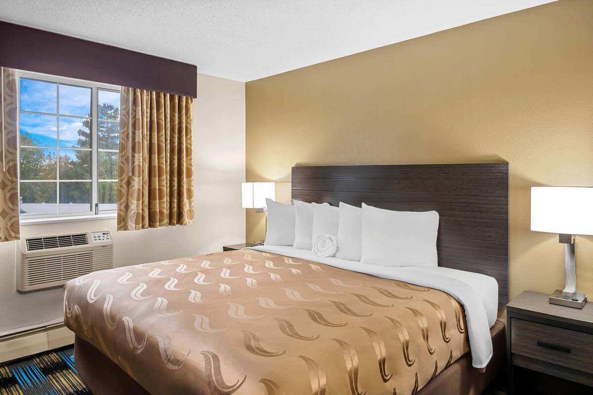 A hotel room with a large bed, two bedside lamps, a window with curtains, and an air conditioning unit.