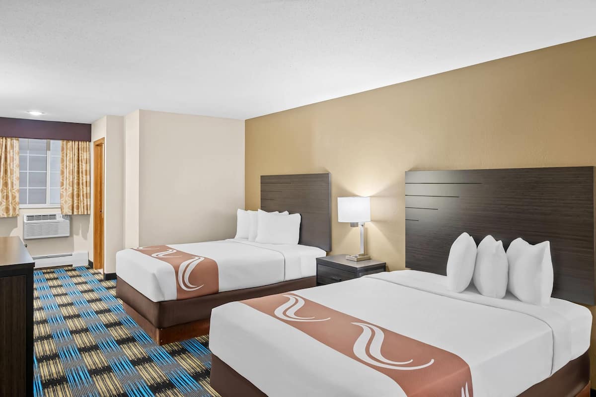 A hotel room with two beds, a desk, a lamp, and a window with curtains.