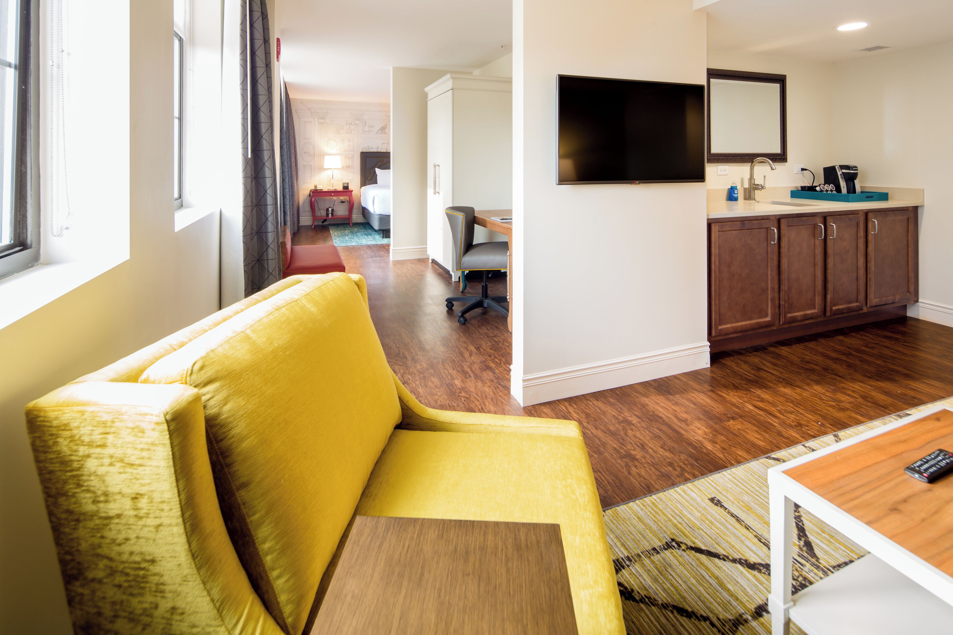 A hotel room with a yellow sofa, a wooden table, a TV mounted on the wall, a kitchen area with cabinets, and a window with curtains.