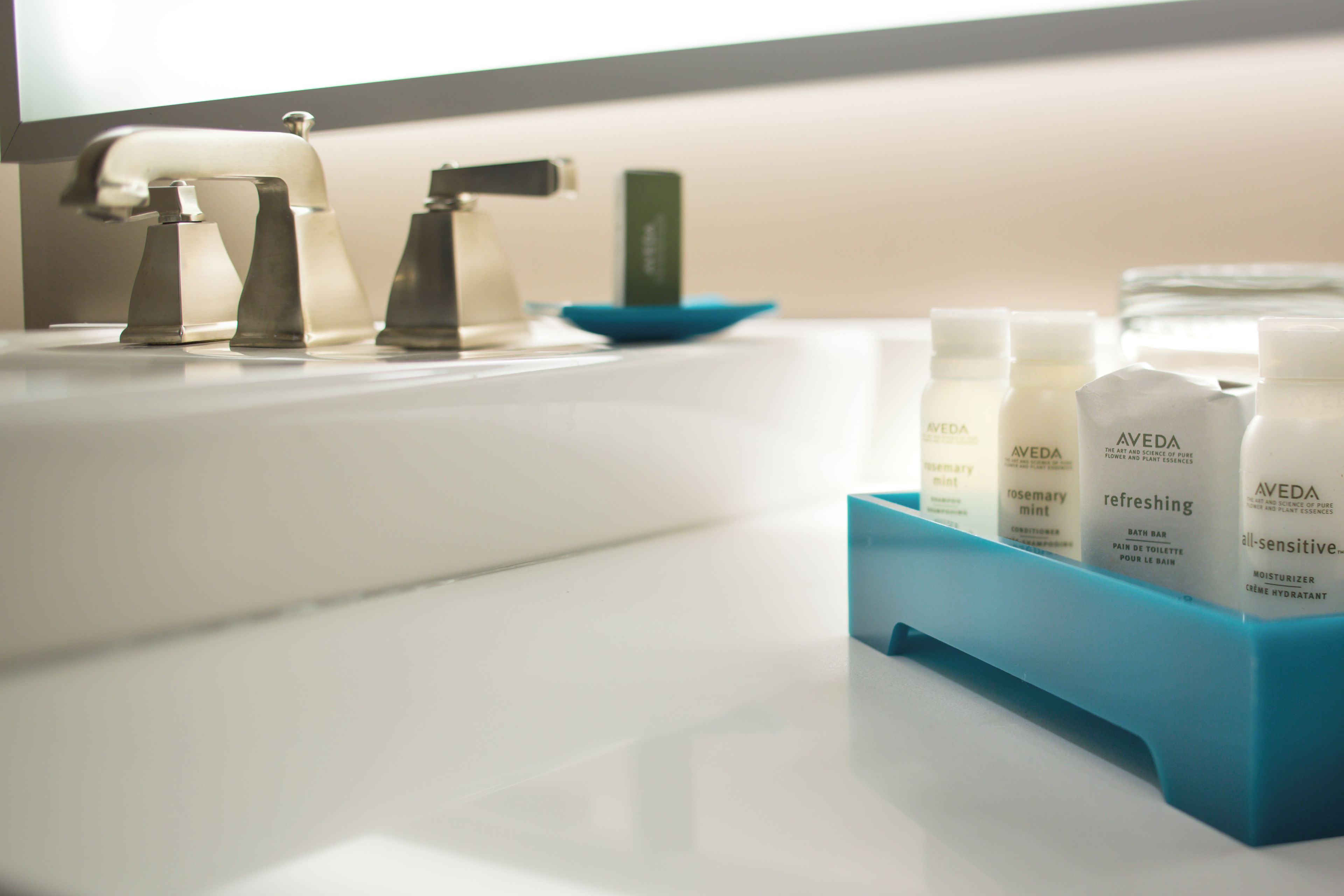 A bathroom with a white sink, two faucets, and a blue soap dish. There are three bottles of Aveda products on the sink.