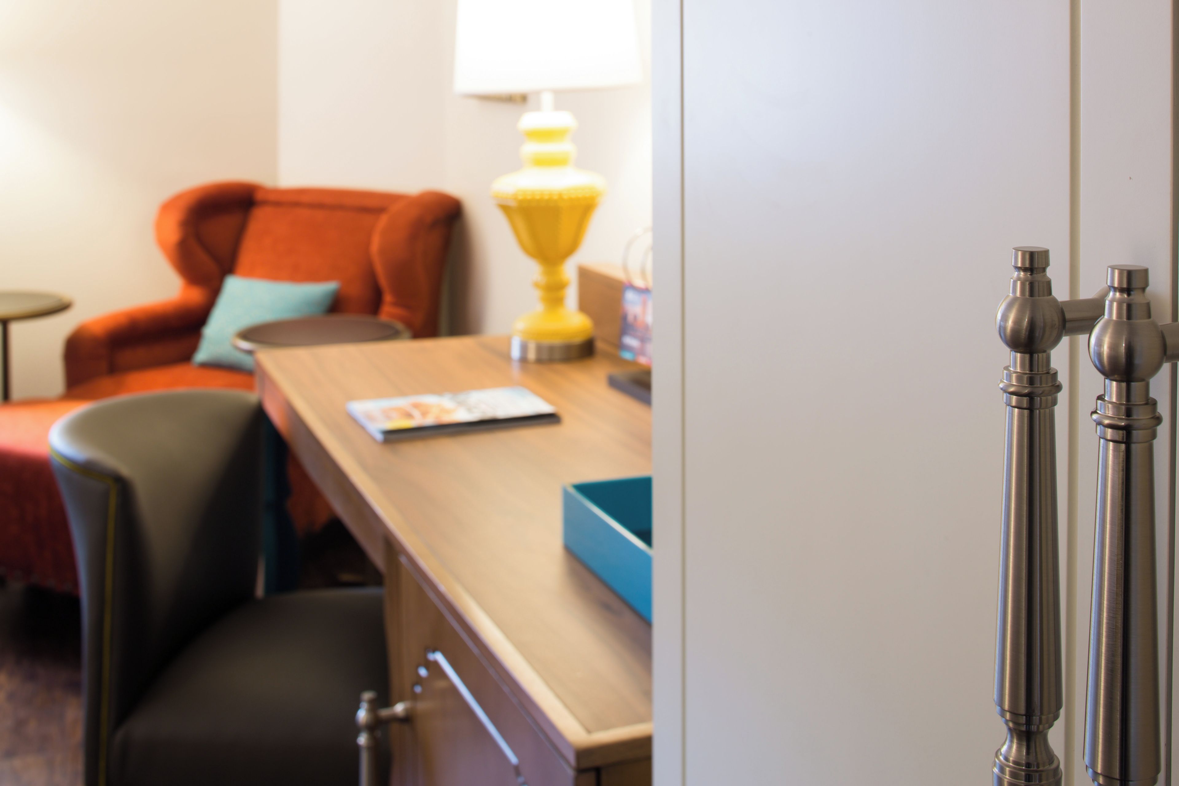 A hotel room with a wooden desk, a chair, a lamp, and a door with a handle.