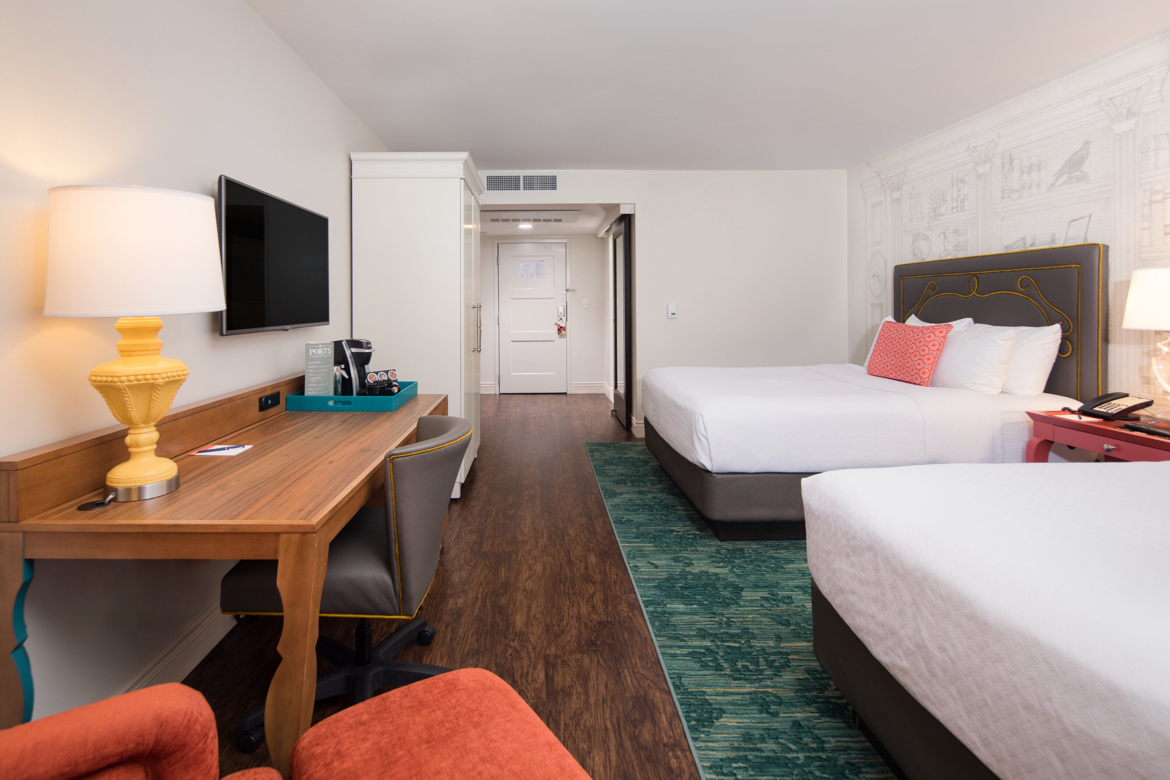 A hotel room with a wooden desk, two beds, a TV, and a small kitchenette.