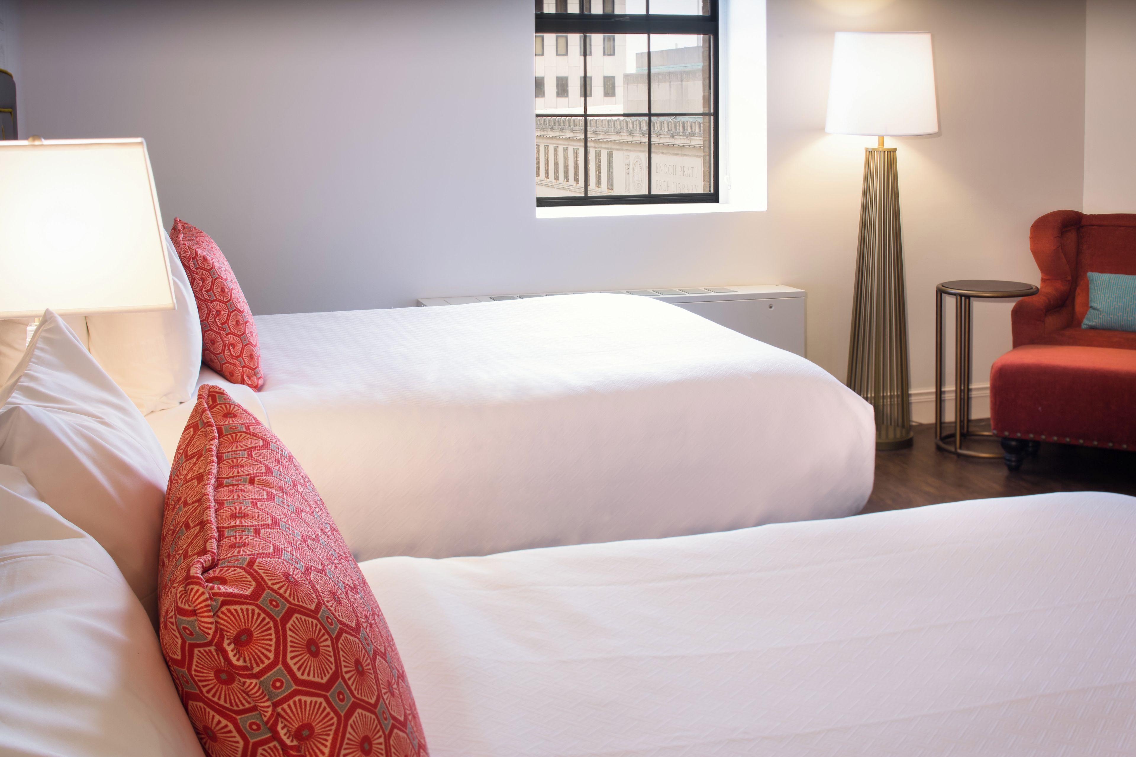 A hotel room with two beds, a red chair, a standing lamp, and a window with a view of a building.