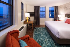 Quarto de hotel com cama, escrivaninha com luminária, cadeira e vista da cidade pela janela, além de uma janela ampla.
