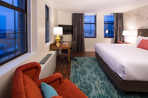 Habitación de hotel con cama, escritorio con lámpara, una silla y vista de la ciudad a través del ventanal.