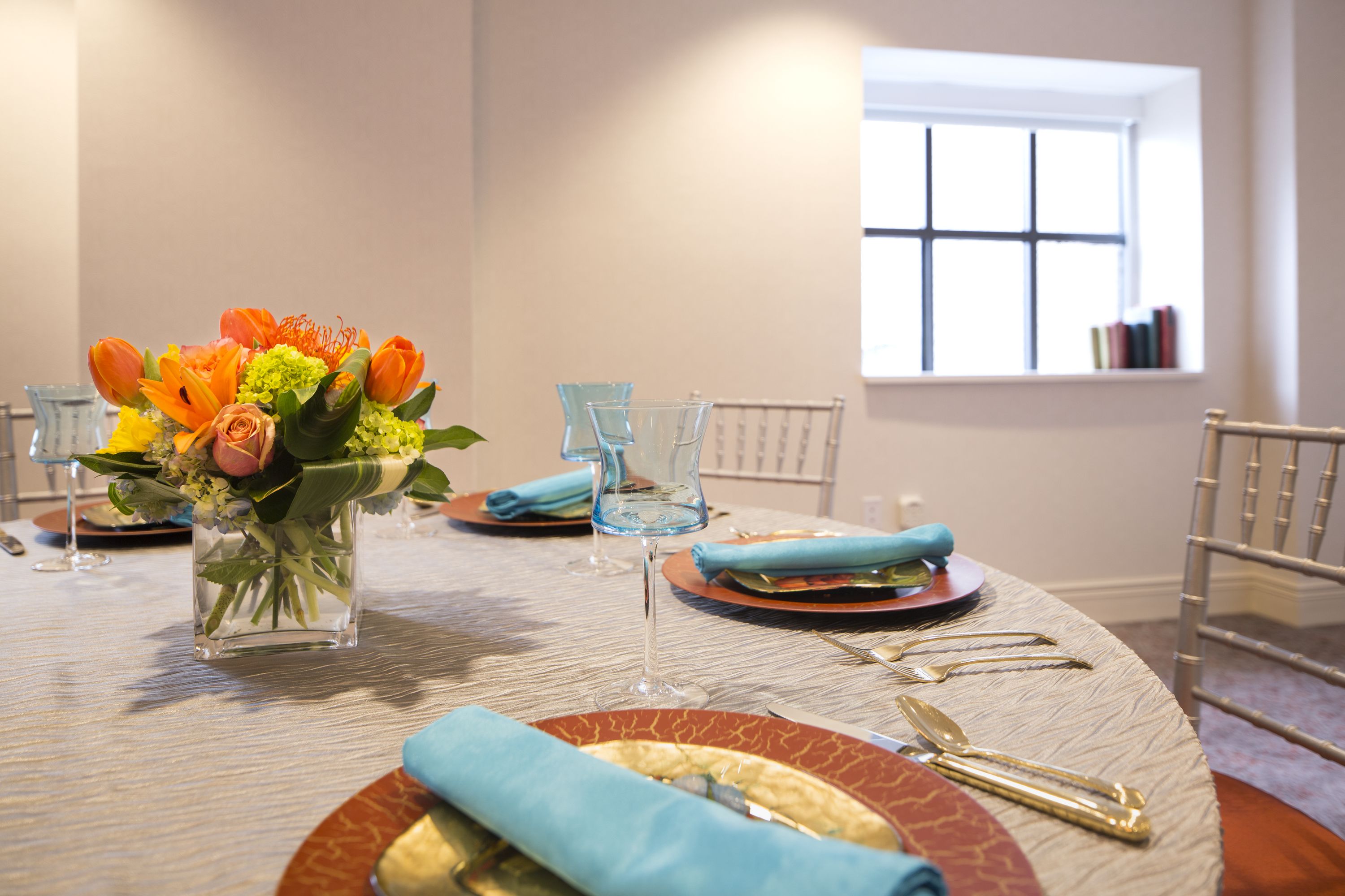 Een eettafel gedekt met blauwe servetten, glazen en een vaas met oranje bloemen.