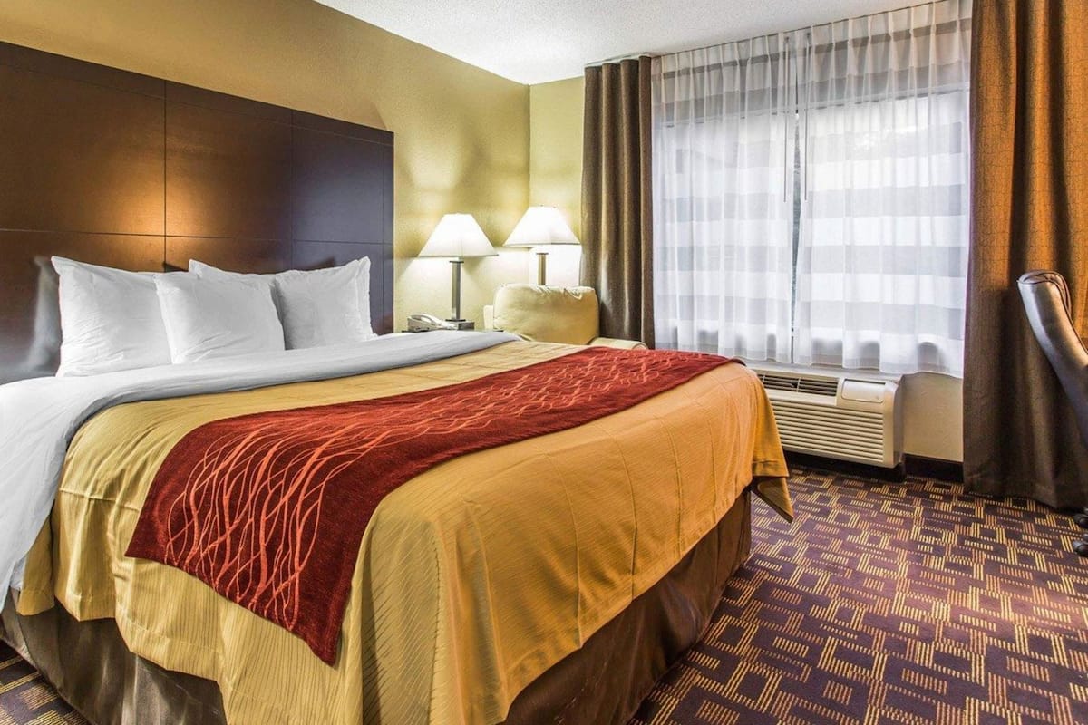 A hotel room with a large bed, two bedside lamps, a window with curtains, and a wall-mounted air conditioning unit.