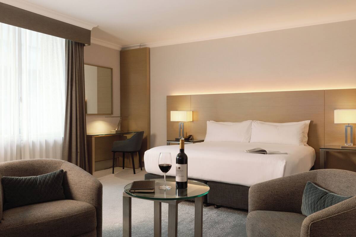 A hotel room with a bed, two bedside lamps, a glass table with a bottle of wine and a glass, and two armchairs.