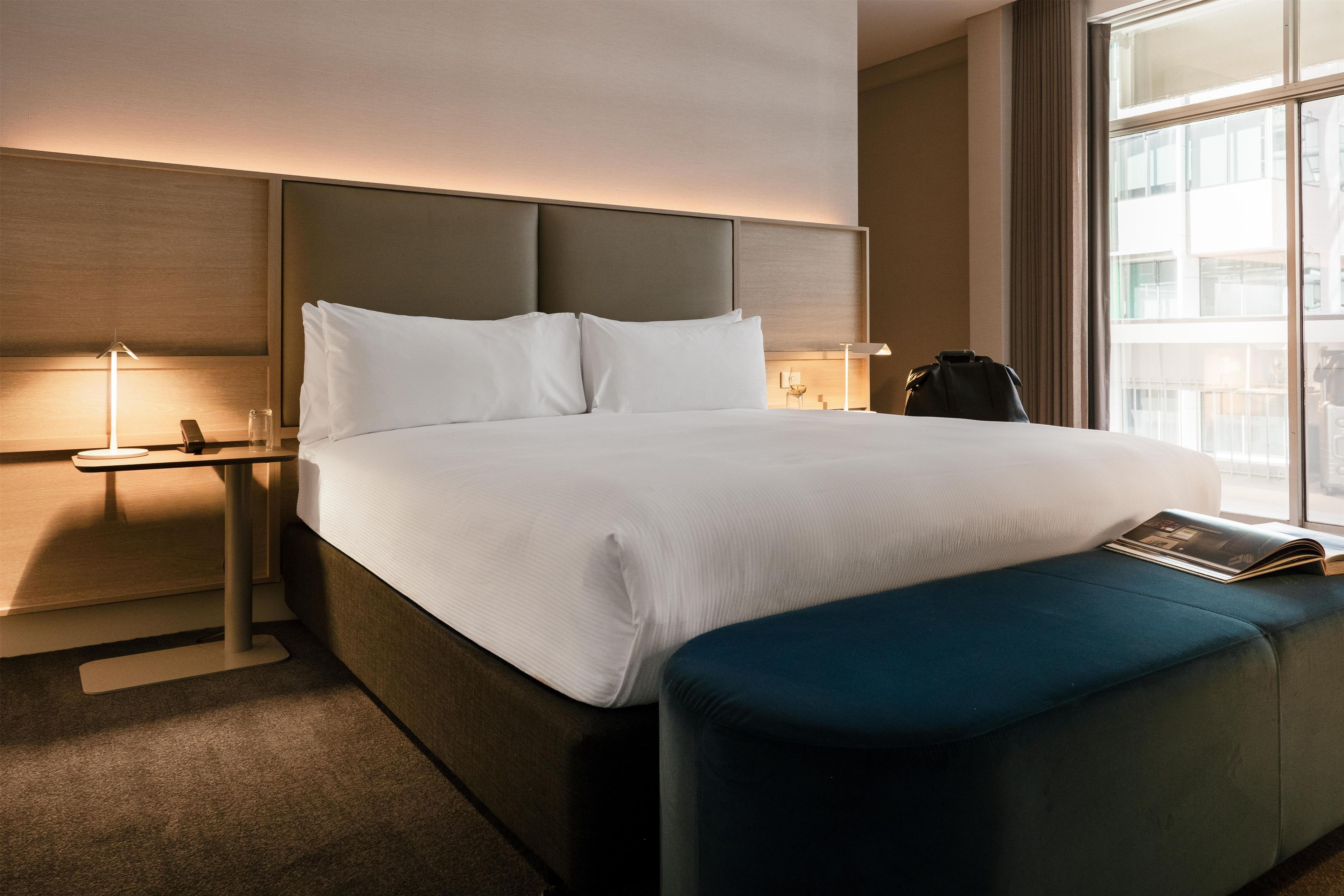 A hotel room with a large bed, a bedside table with a lamp, a window with a view, and a blue ottoman with a magazine.