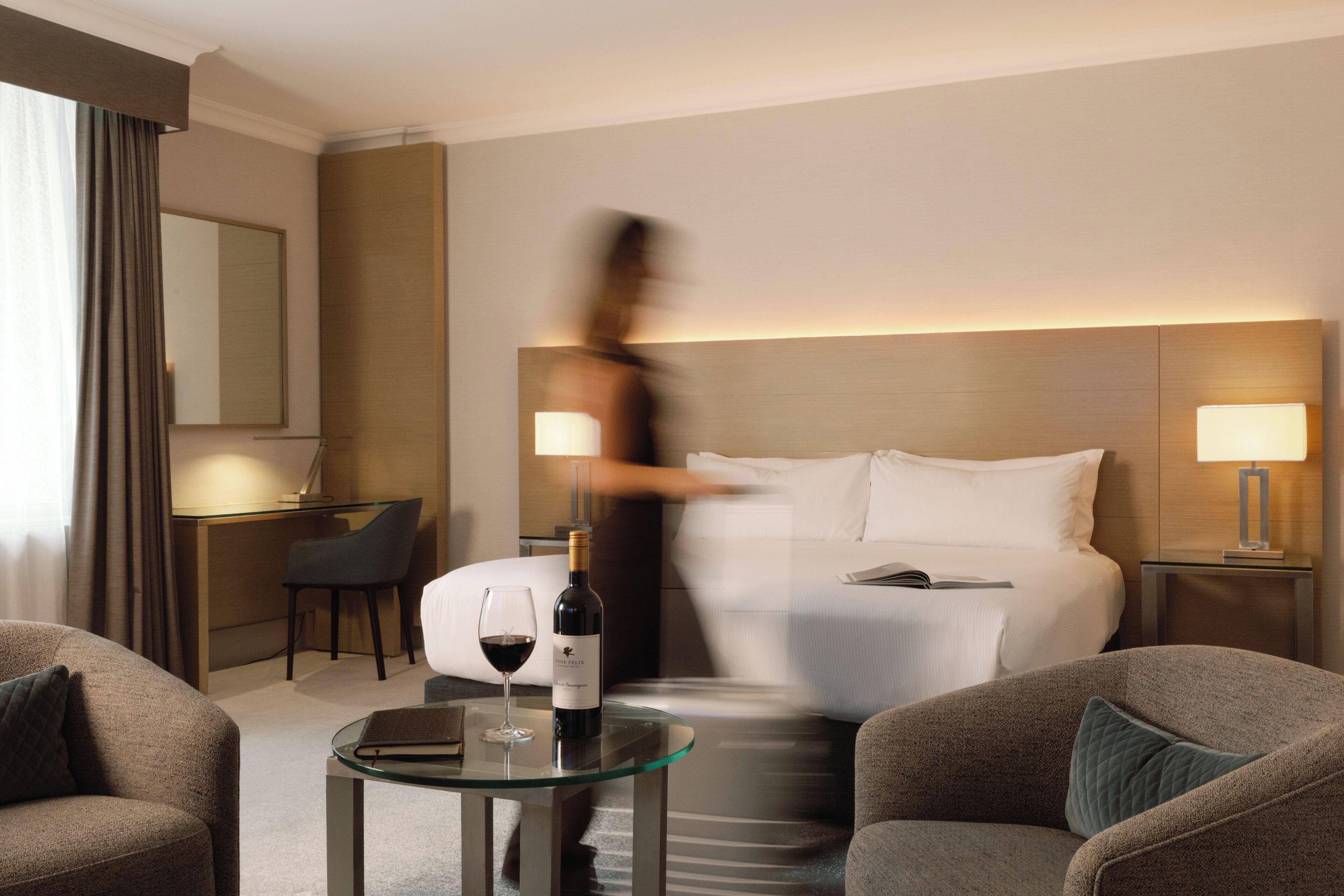 Una habitación de hotel con una cama, dos lámparas de noche, una mesita de cristal con una botella de vino y una copa, una silla y un escritorio con una silla.