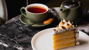 A slice of layered cake on a white plate, a cup of tea, and a teapot on a marble table.