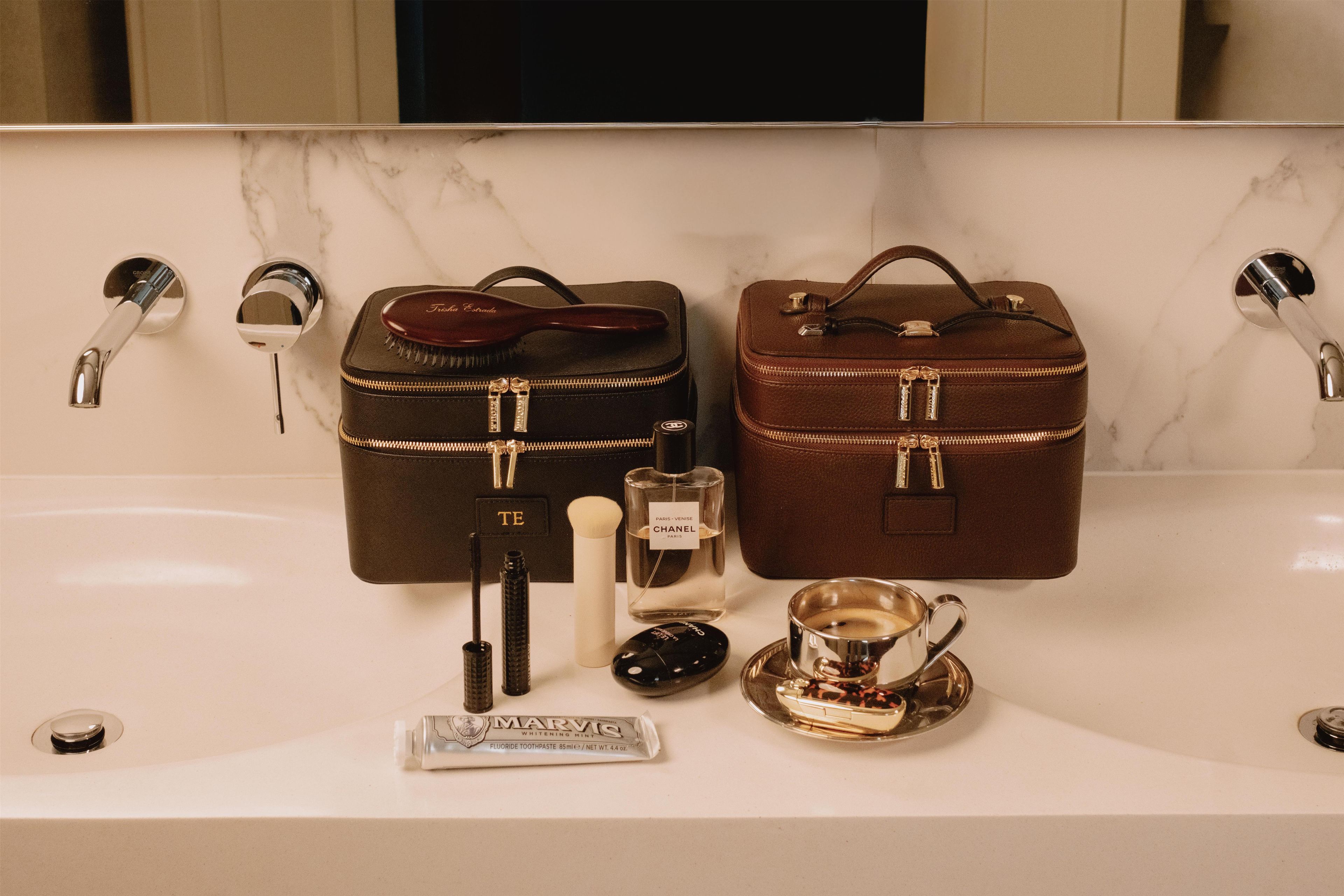 Un tocador de baño con dos bolsas de cuero marrón, una botella de perfume, un pincel de maquillaje, un tubo de pasta de dientes y una taza de café.