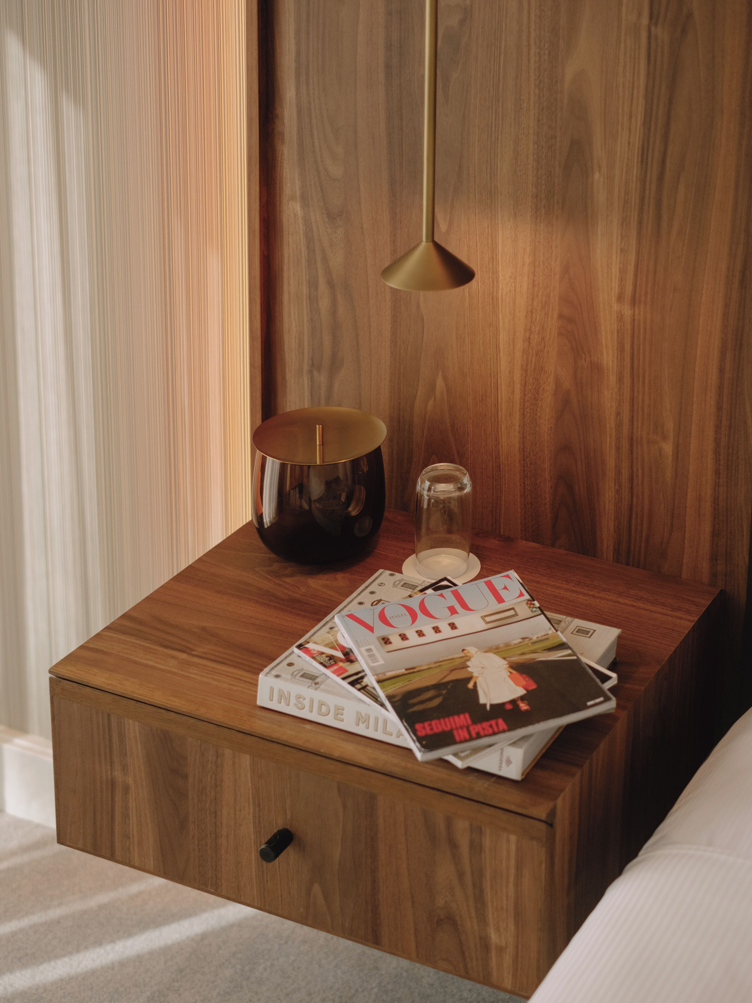 A wooden bedside table with a lamp, a candle, and magazines.