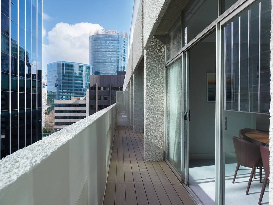 A balcony with a view of modern buildings, a glass door leading to a room with a table and chairs, and a wooden floor.