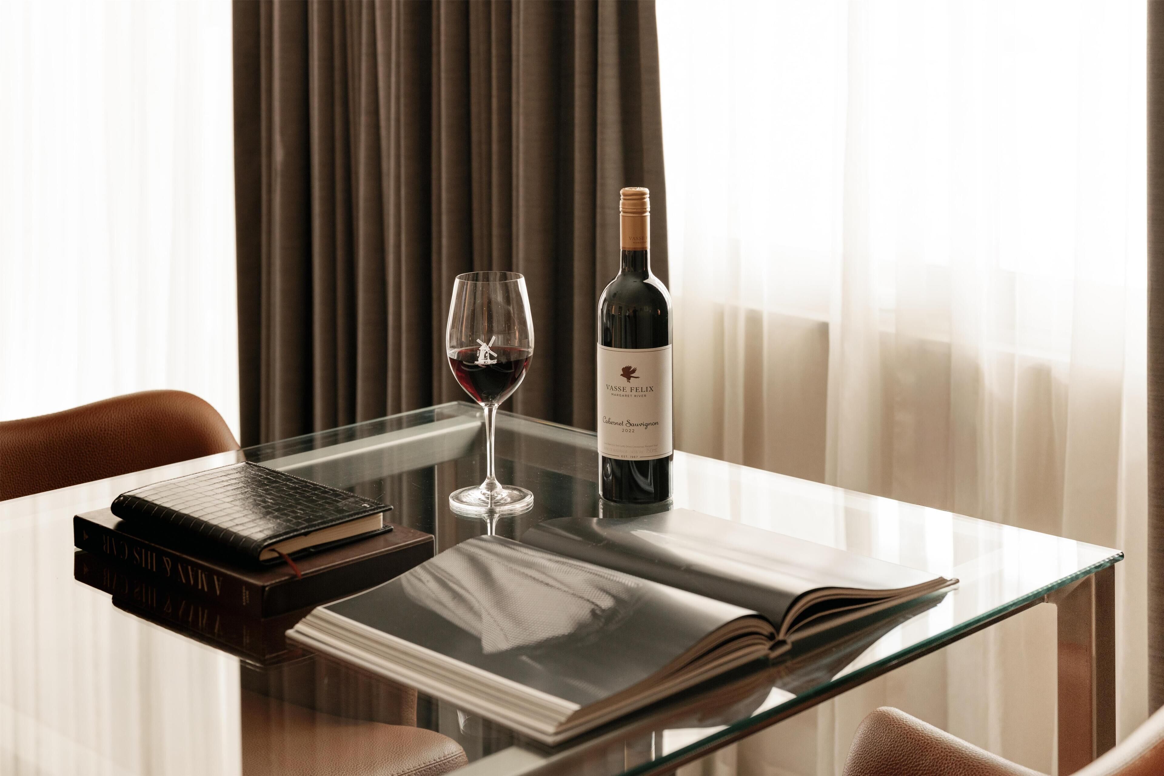 Une table en verre avec une bouteille de vin, un verre de vin rouge et des livres.