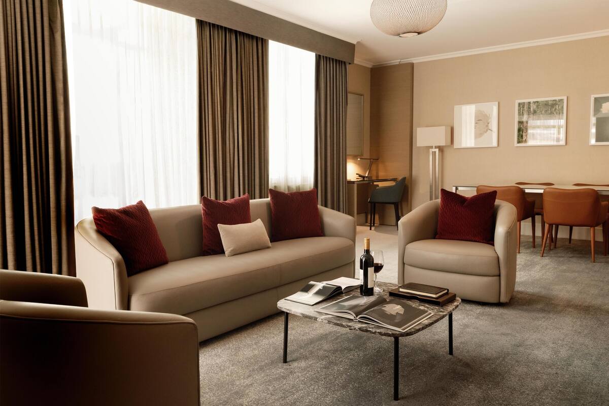 A hotel room with a sofa, armchairs, a coffee table with books and a bottle, and a dining area with chairs and a table.