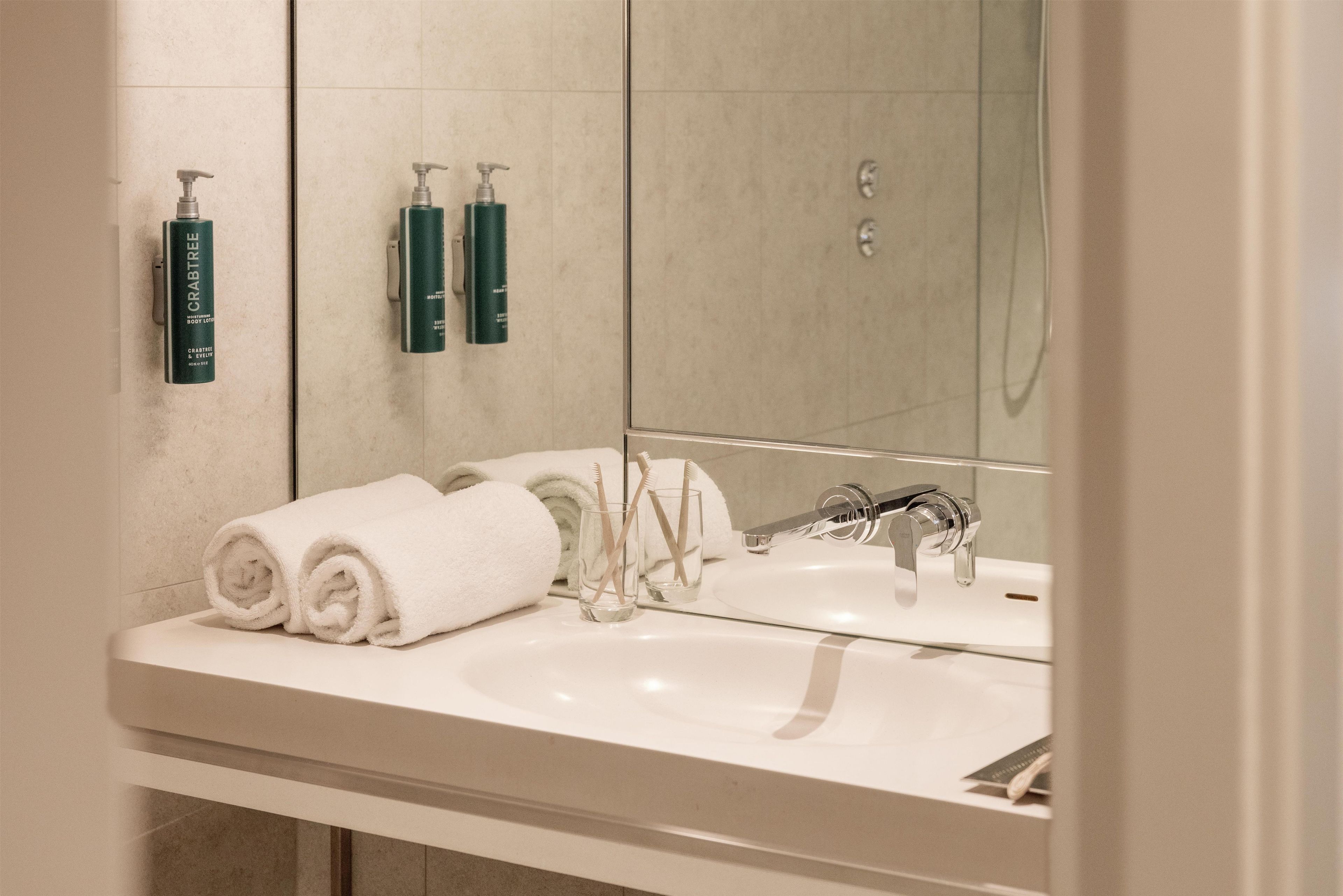 Un baño con un lavabo blanco, dos cepillos de dientes, un vaso de agua y dos toallas enrolladas.