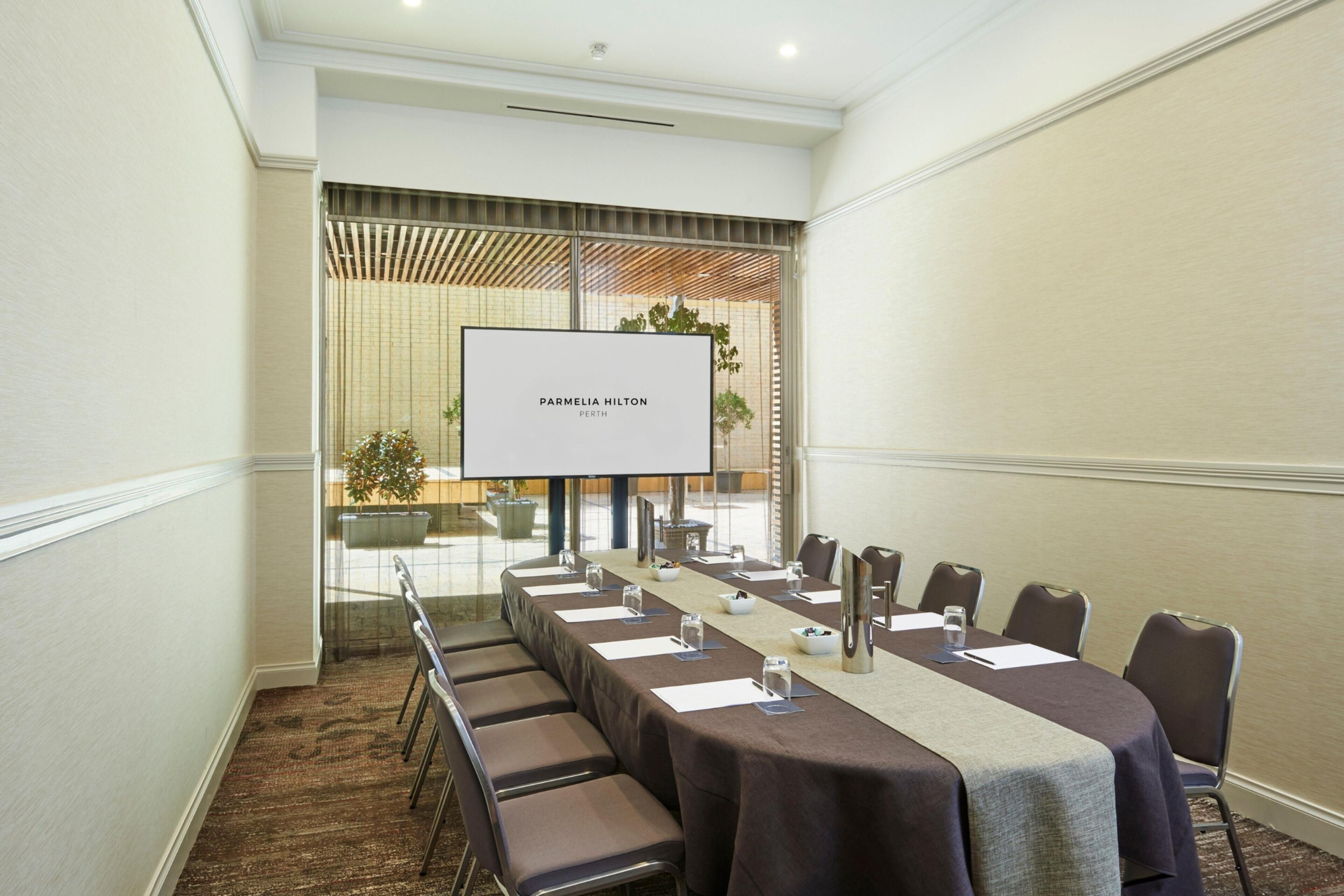 Een vergaderruimte met een lange tafel, stoelen, een scherm voor een projector en een plant.