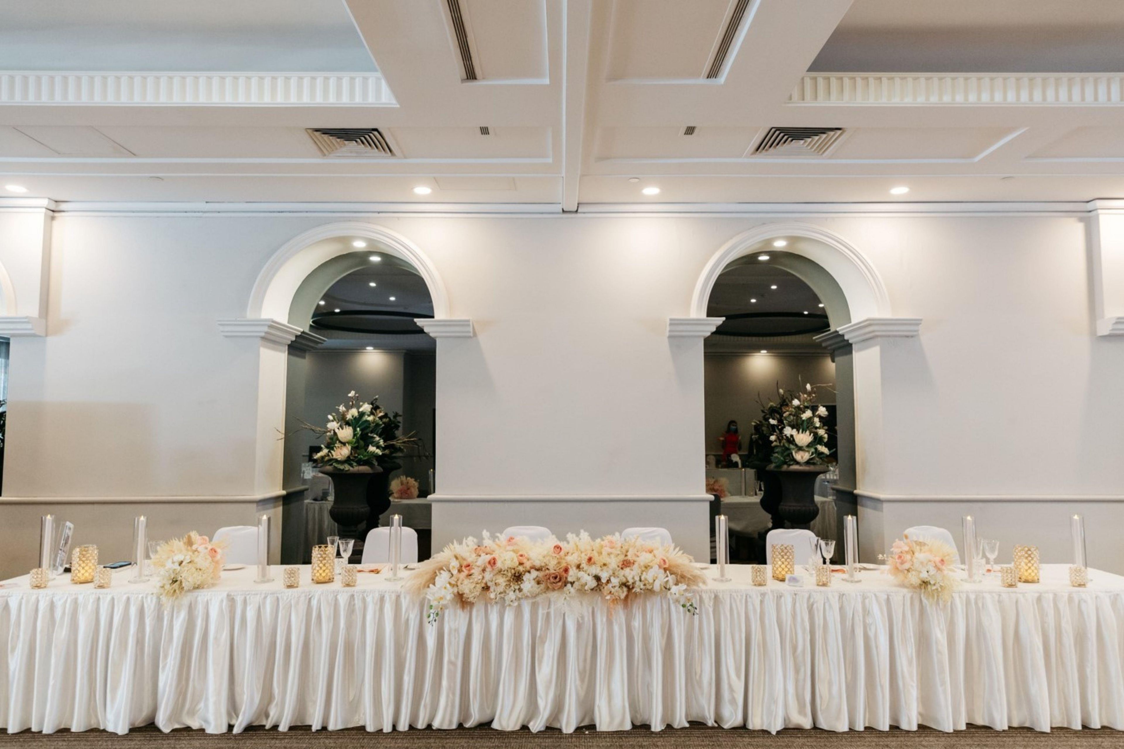 Une salle de banquet avec une longue table dressée pour un événement formel, flanquée de deux arcades ornées de compositions florales.