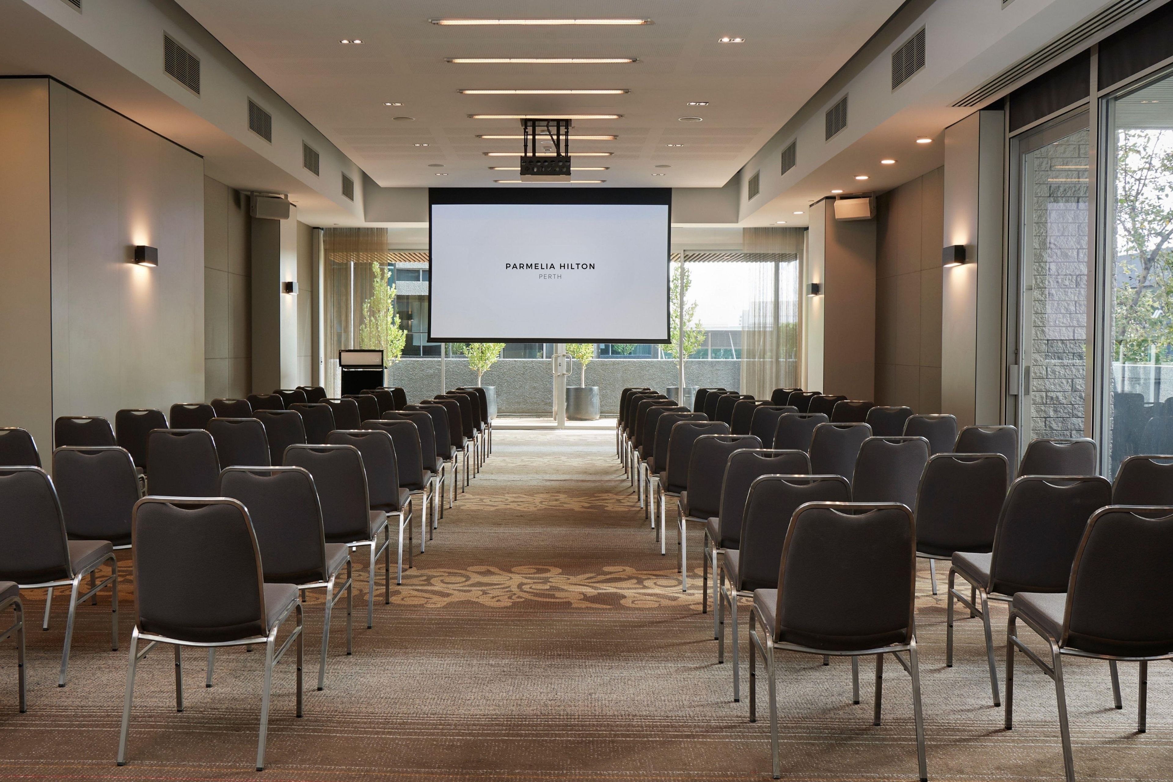 Ein Konferenzraum mit Reihen von Stühlen, einem großen Bildschirm, der 'PARMELIA HILTON PERTH' zeigt, und einem Blick ins Freie durch große Fenster.