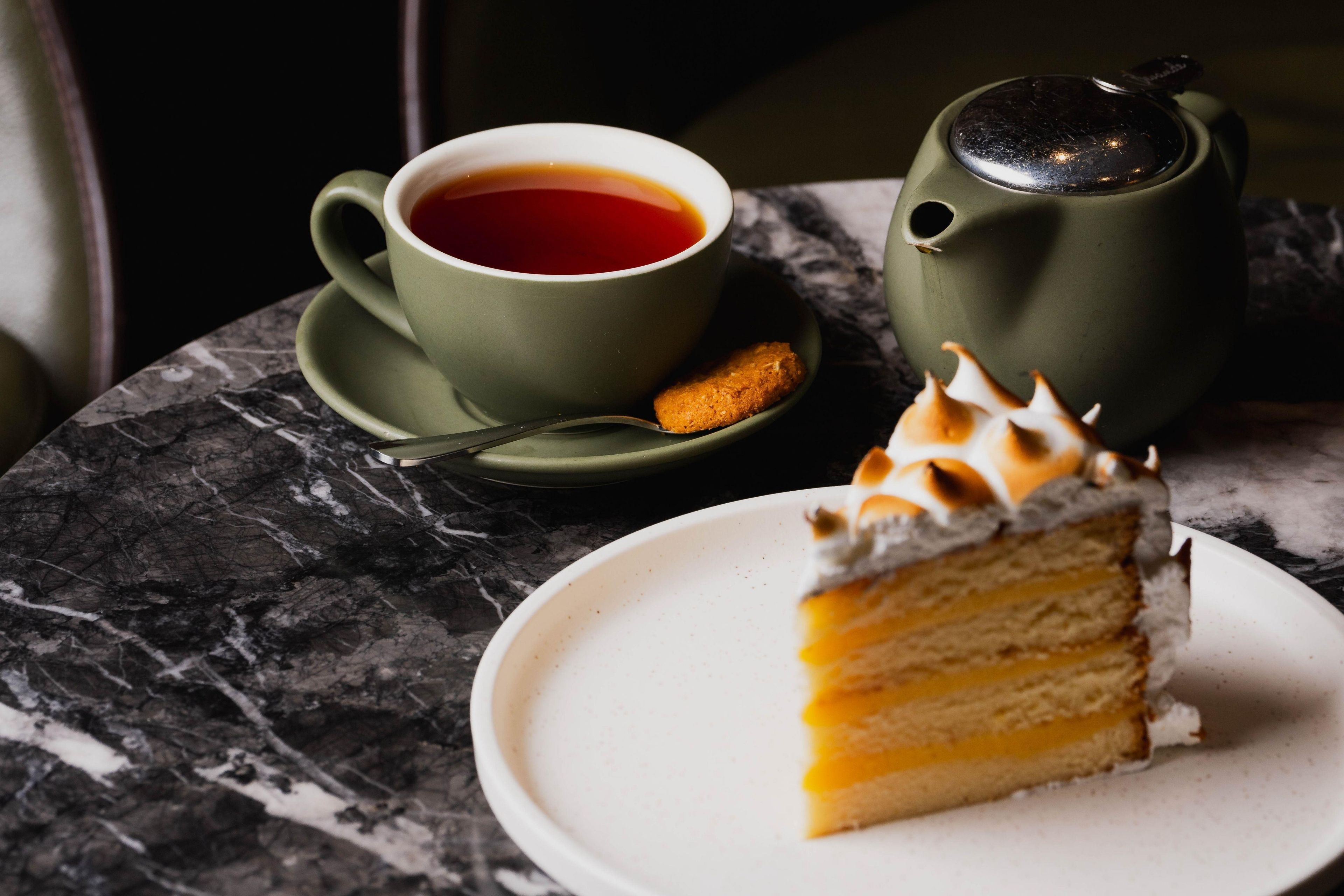 A slice of layered cake on a white plate, a cup of tea on a saucer, and a teapot on a table.