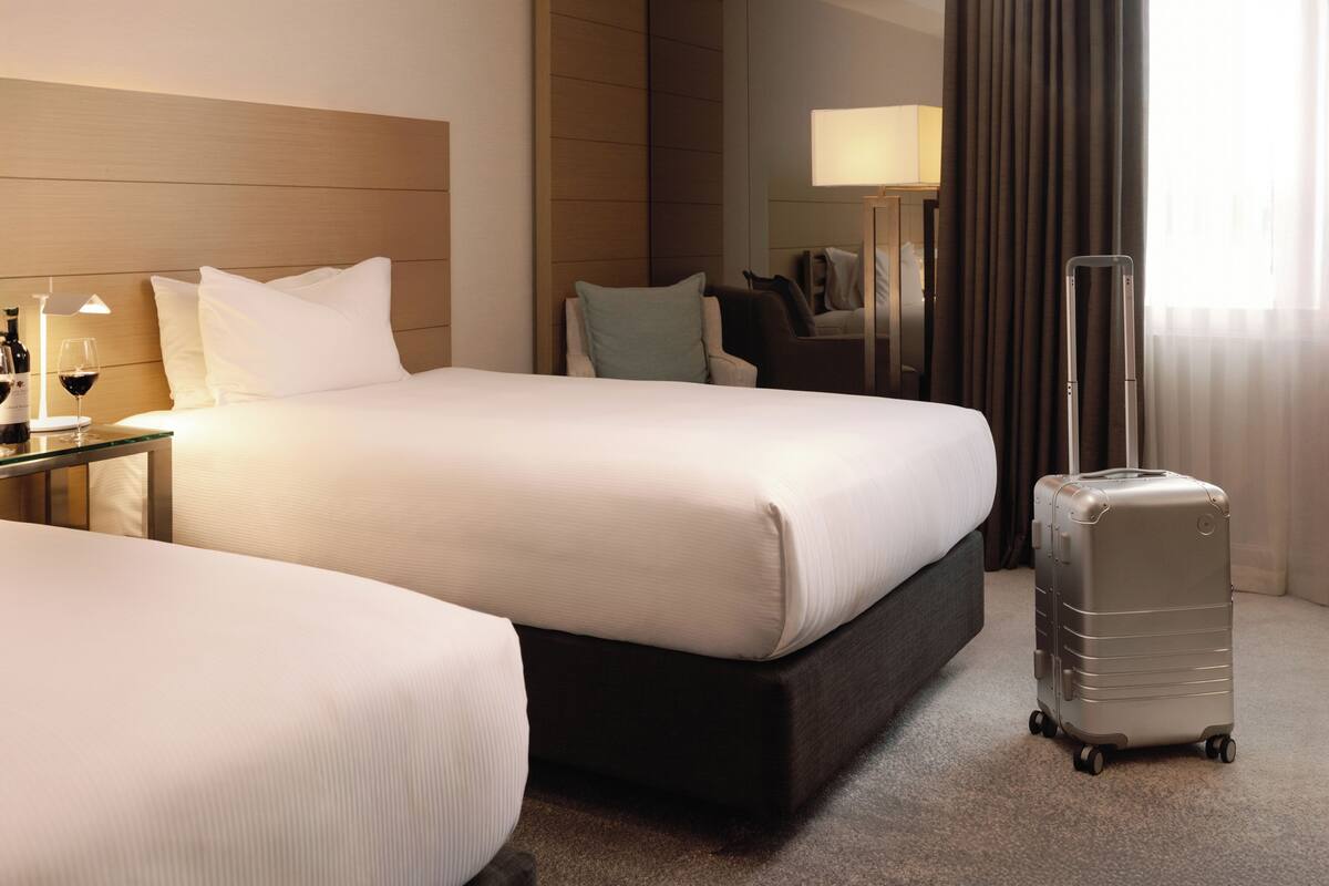 A hotel room with two beds, a nightstand with a lamp, a suitcase, and a view of the outdoors.