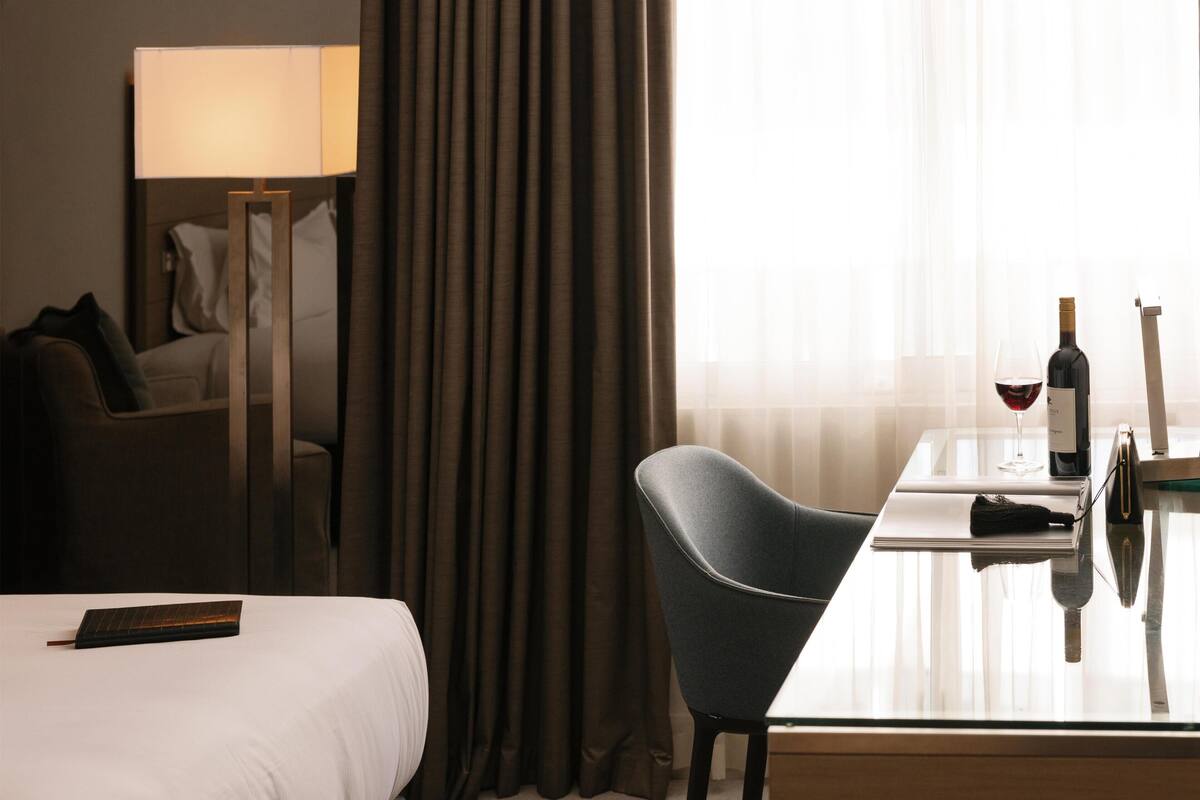 A hotel room with a bed, a desk with a chair, a lamp, a bottle of wine, and a glass.