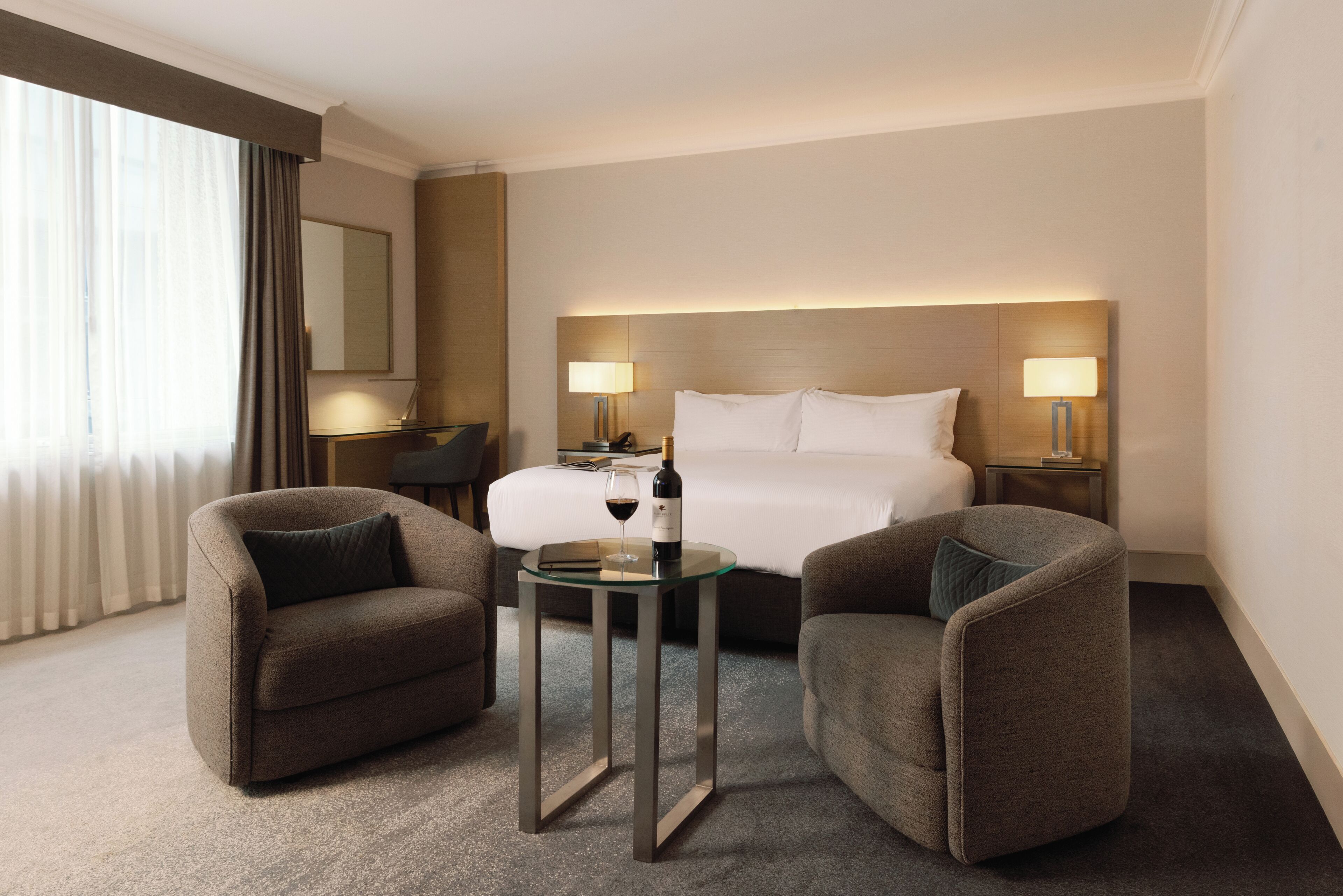 A hotel room with a bed, two armchairs, a small table with a bottle and glass, and a nightstand with a lamp.