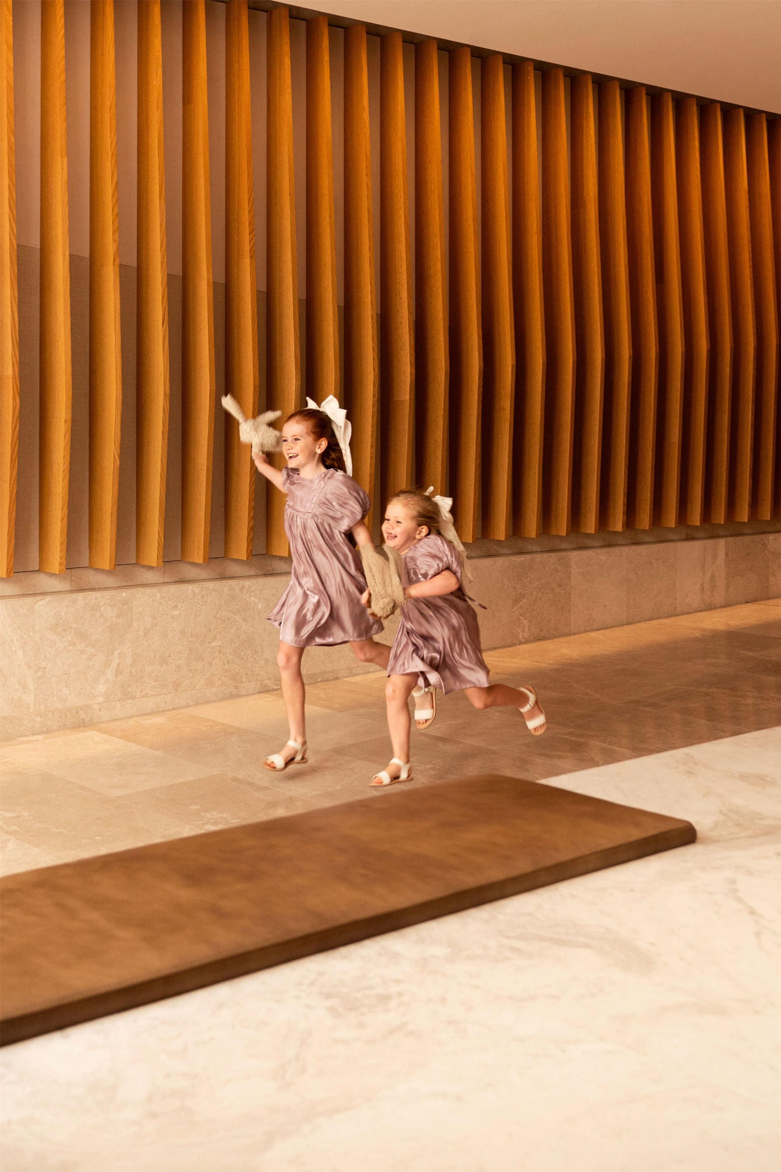 Two children in a hallway with a wooden slatted wall and a marble floor.