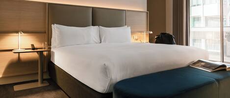 A hotel room with a large bed, a bedside table with a lamp, a window with a view, and a blue ottoman.