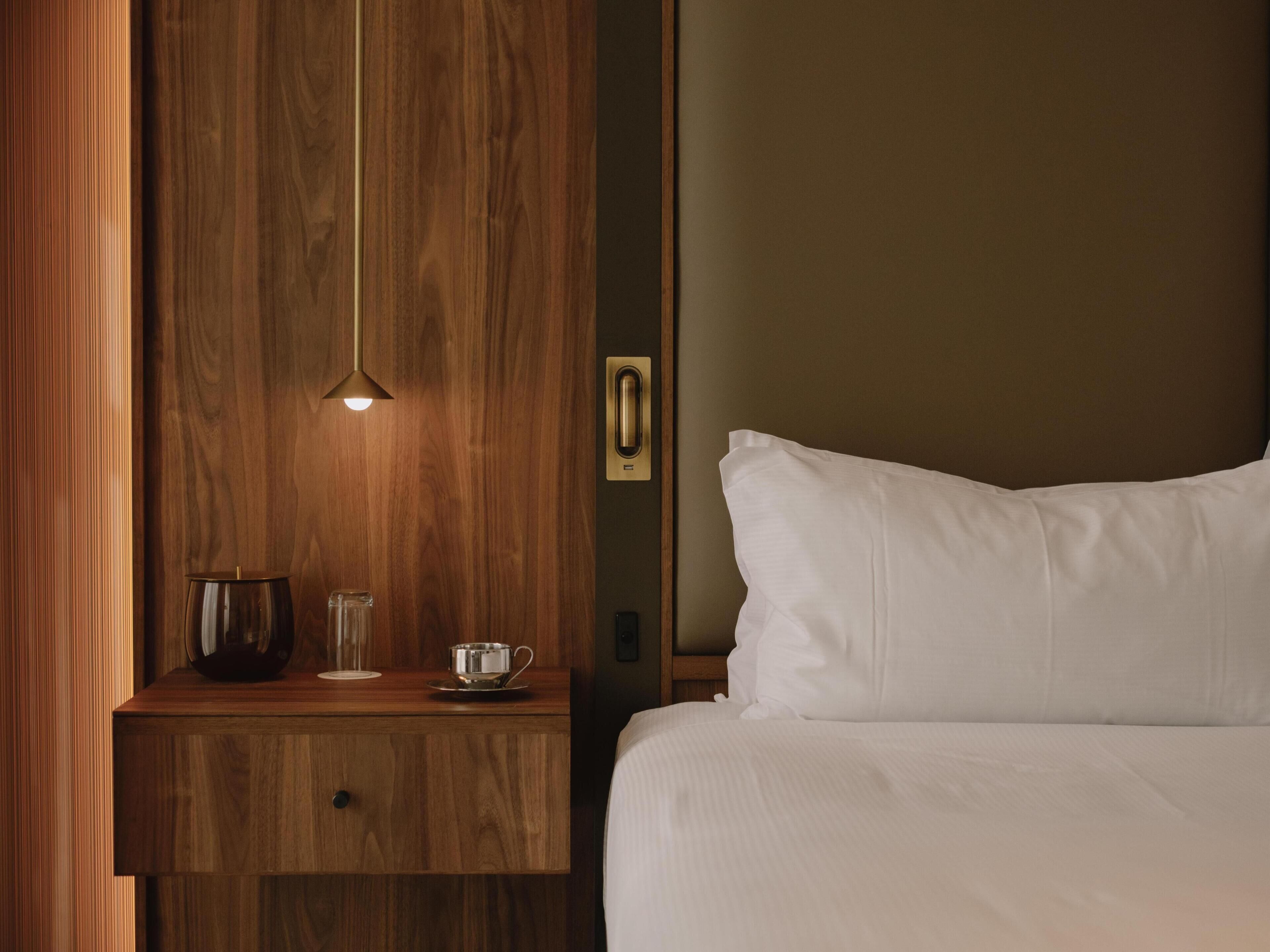 A neatly made bed with white linens, a wooden nightstand with a cup and saucer, and a pendant light.