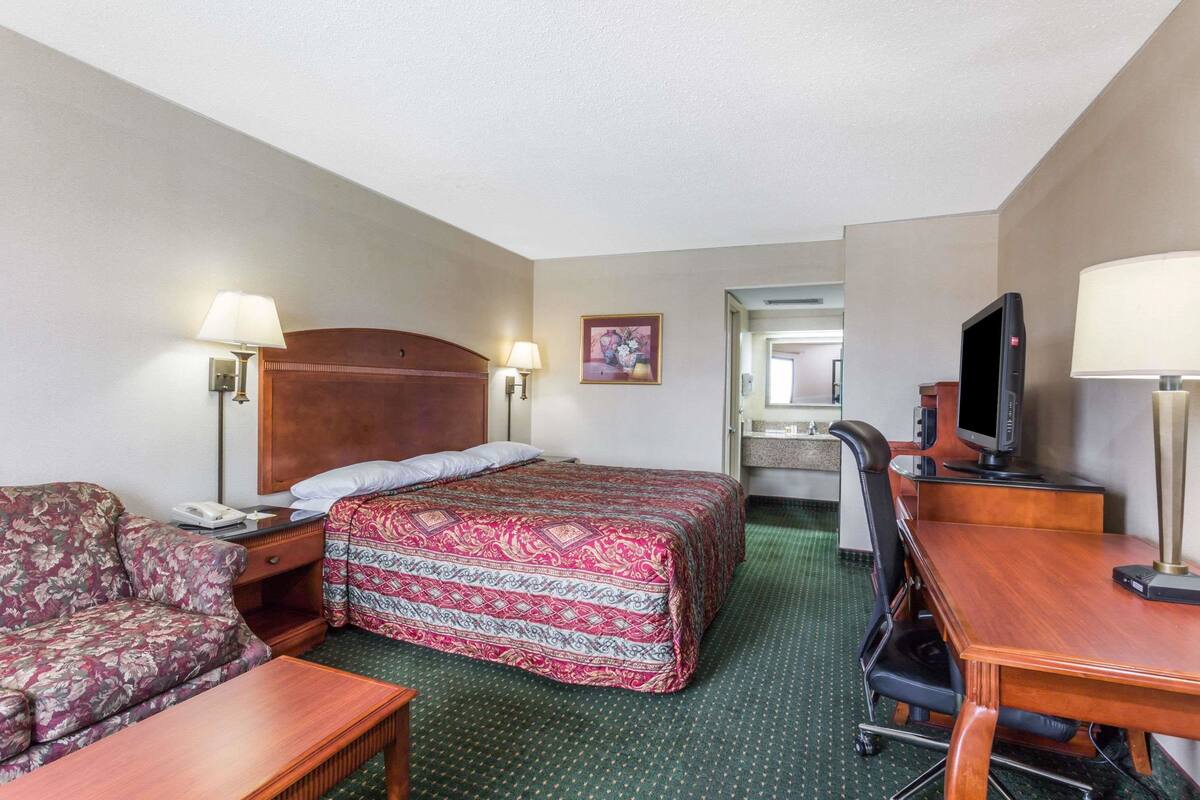 A hotel room with a bed, a sofa, a desk, a television, and a bathroom visible in the background.