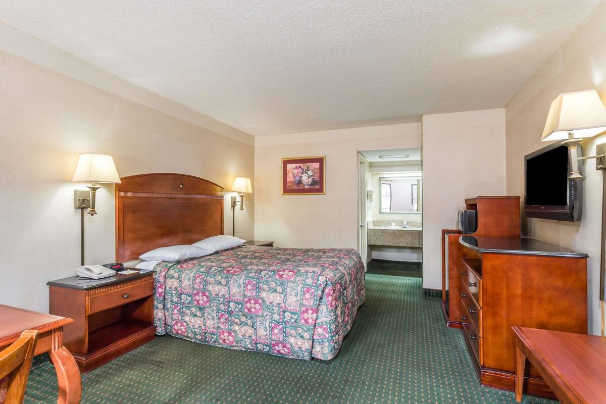 A hotel room with a bed, a desk, a dresser, a television, and a bathroom visible through an open door.
