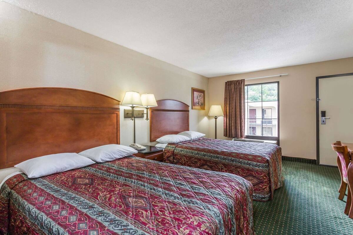 A hotel room with two beds, a nightstand, a lamp, a window with curtains, and a door.