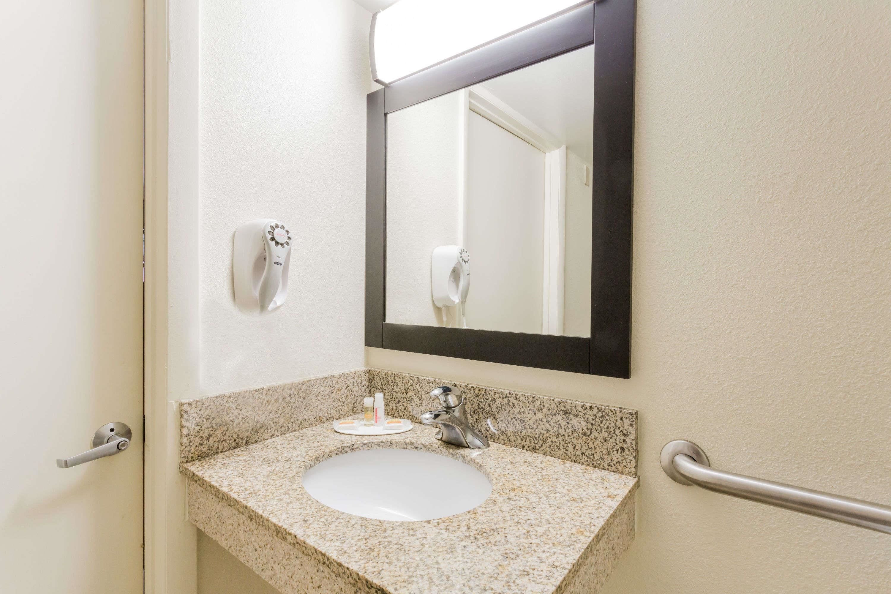 A bathroom with a granite countertop sink, a mirror, a hand dryer, and a hand soap dispenser.