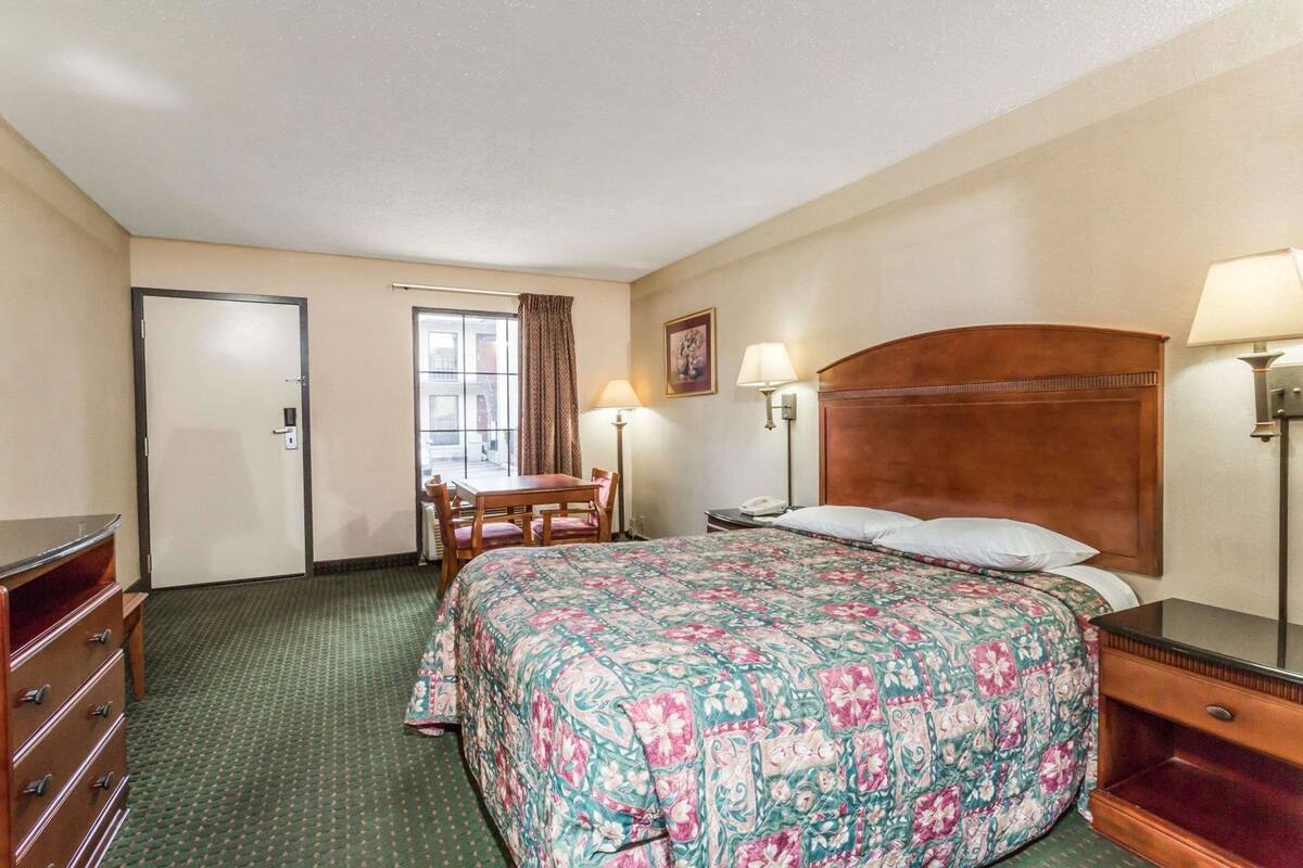 A hotel room with a bed, a desk, a chair, a dresser, a window with curtains, and two bedside lamps.