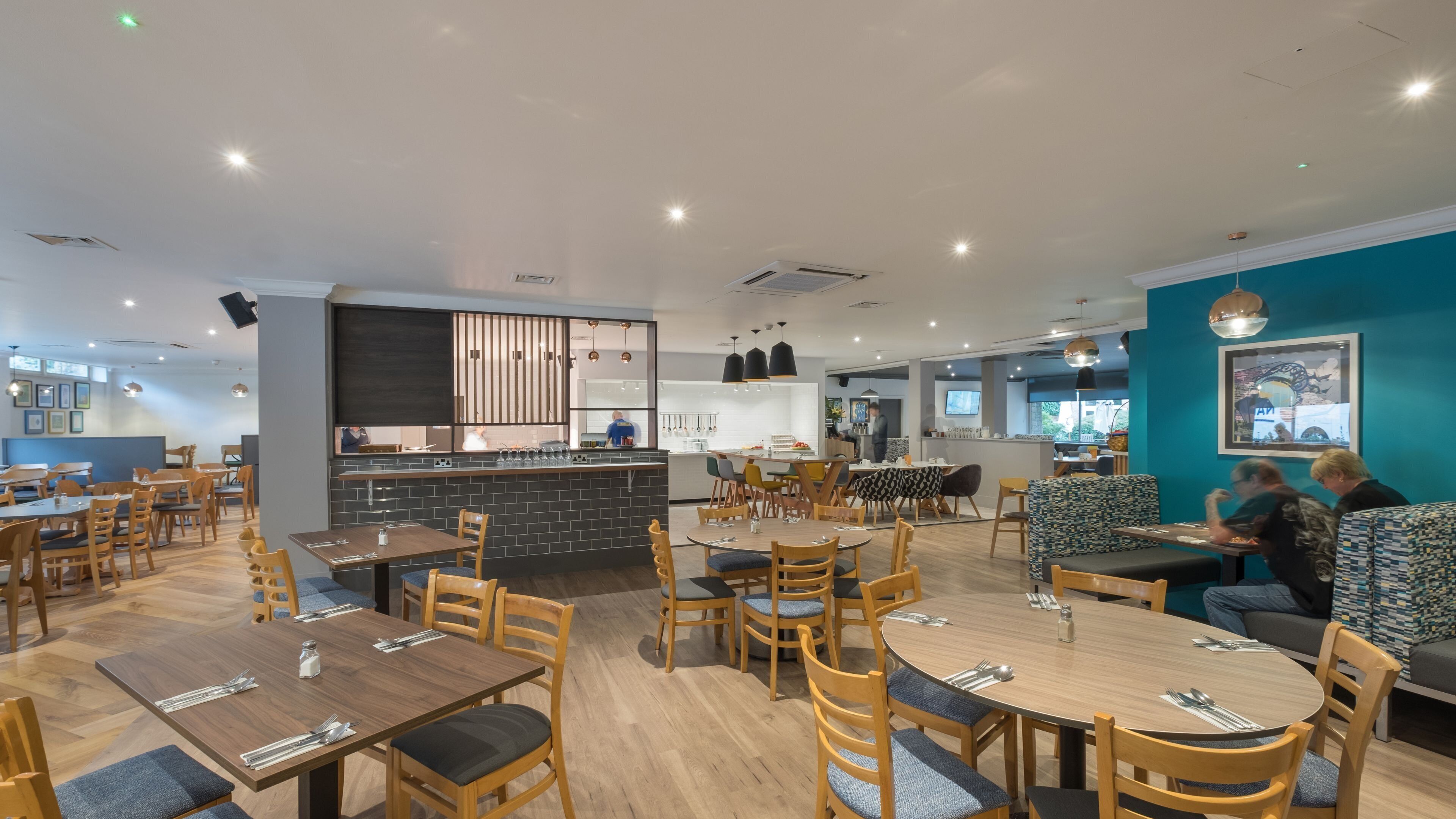 Un restaurante moderno con mesas y sillas de madera, una zona de bar y un área de estar con cojines estampados.