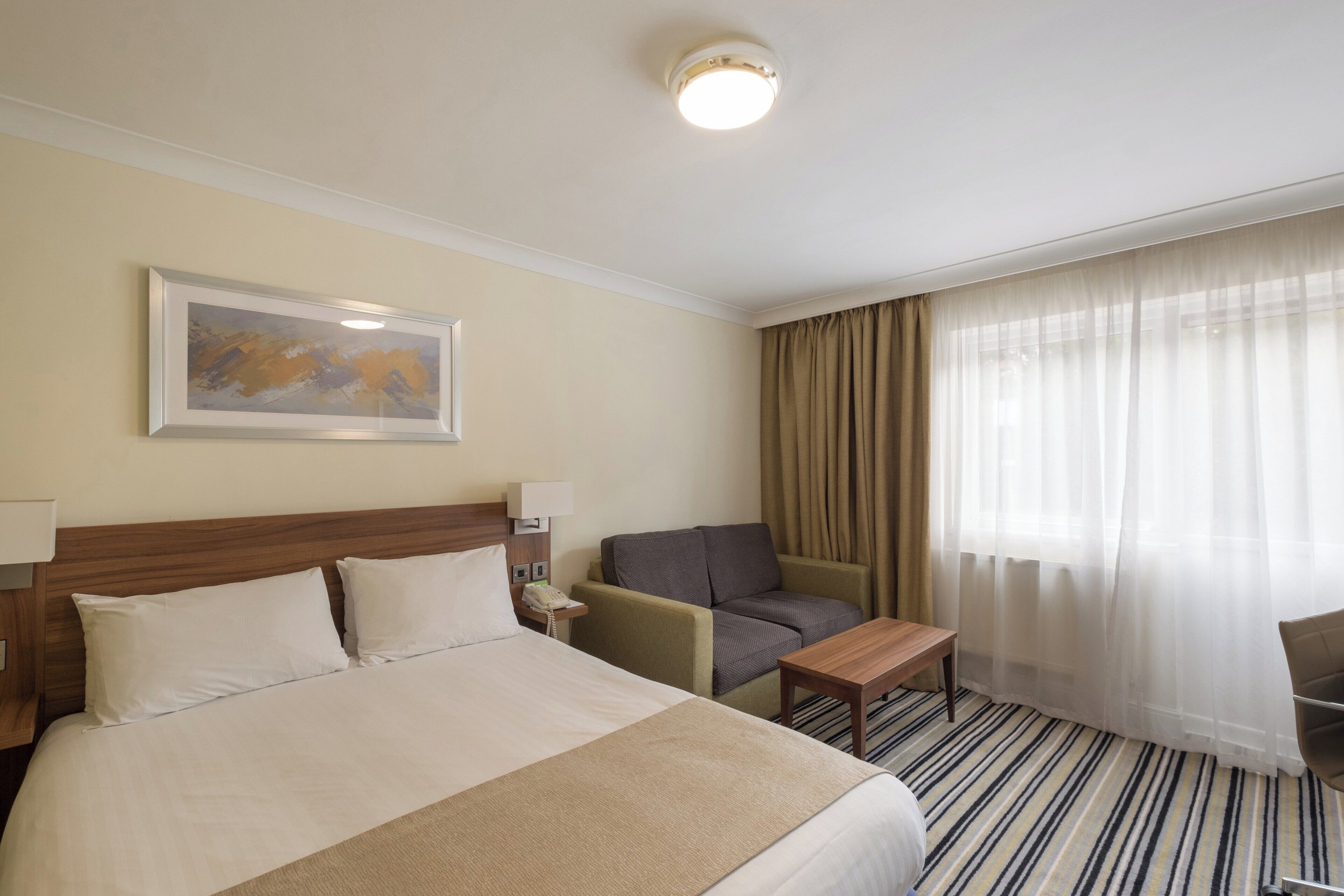 A hotel room with a bed, a desk, a chair, a painting on the wall, and a window with curtains.