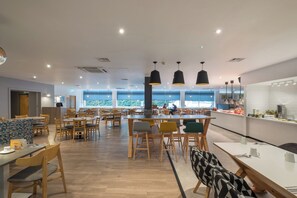 A modern dining area with wooden tables and chairs, pendant lighting, and large windows offering a view of the outdoors. - Holiday Inn Swindon by IHG (Swindon)
