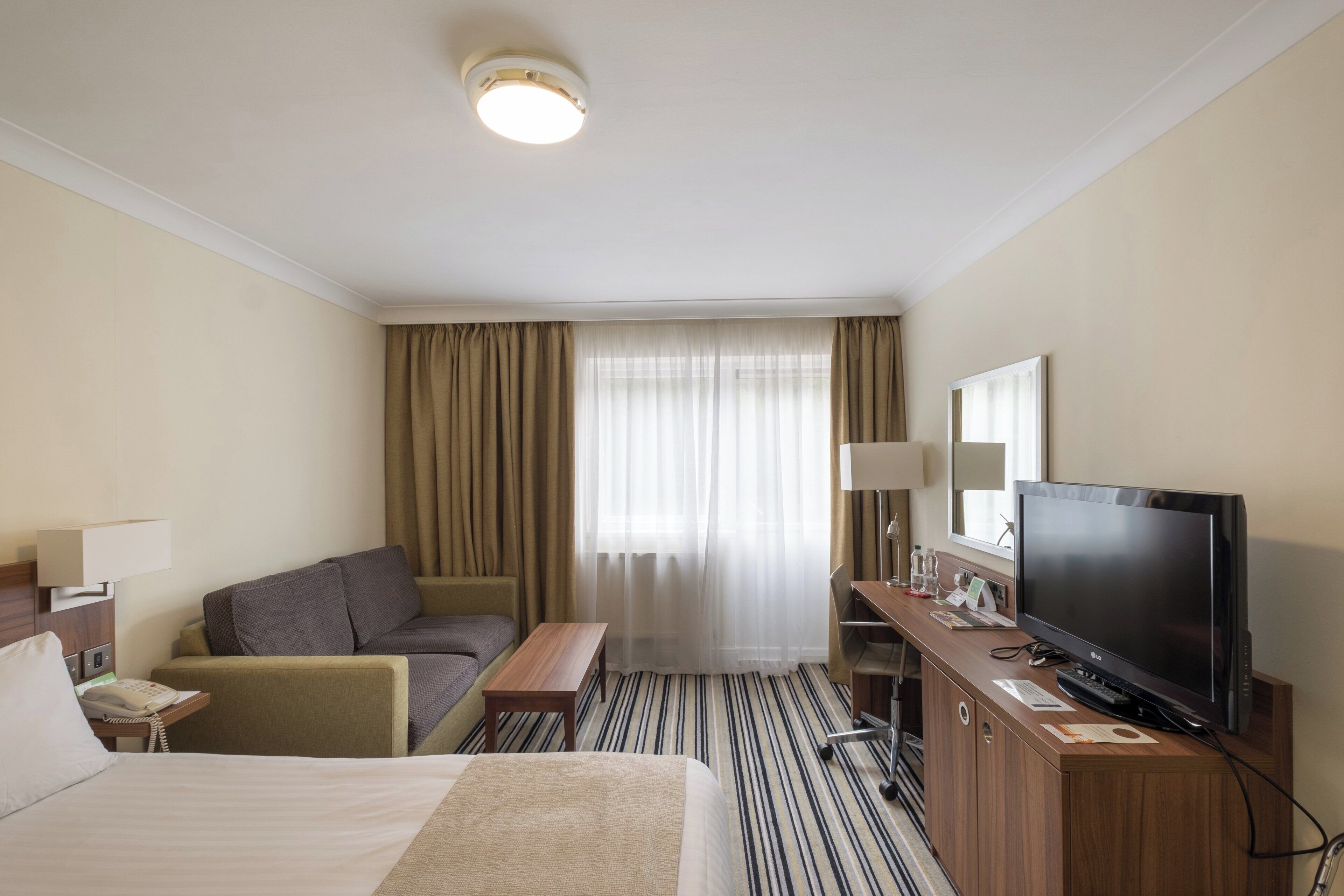 A hotel room with a bed, a sofa, a TV, a desk, and a window with curtains.
