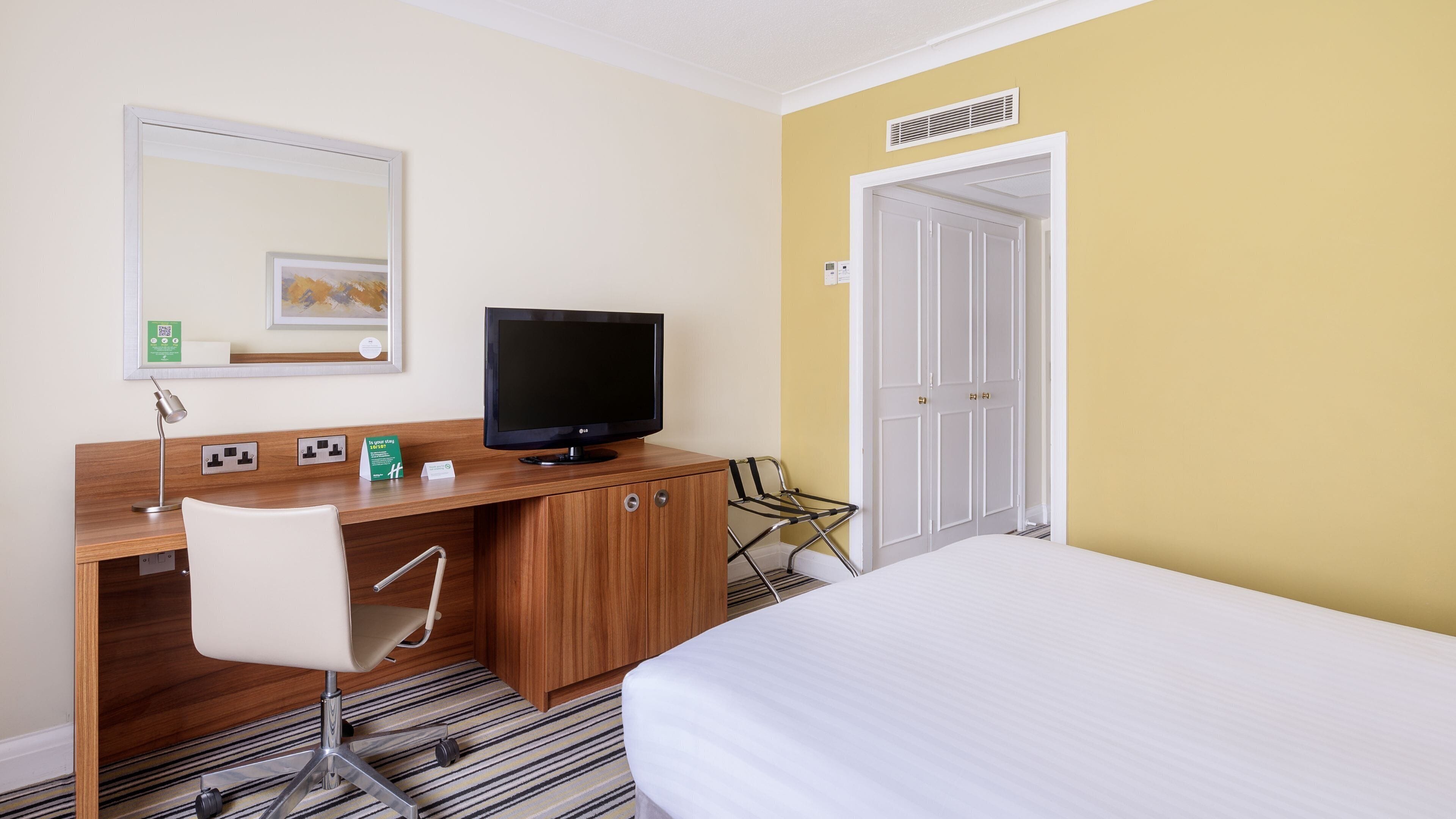 A hotel room with a wooden desk, a television, a white chair, a bed, and a door with a white frame.
