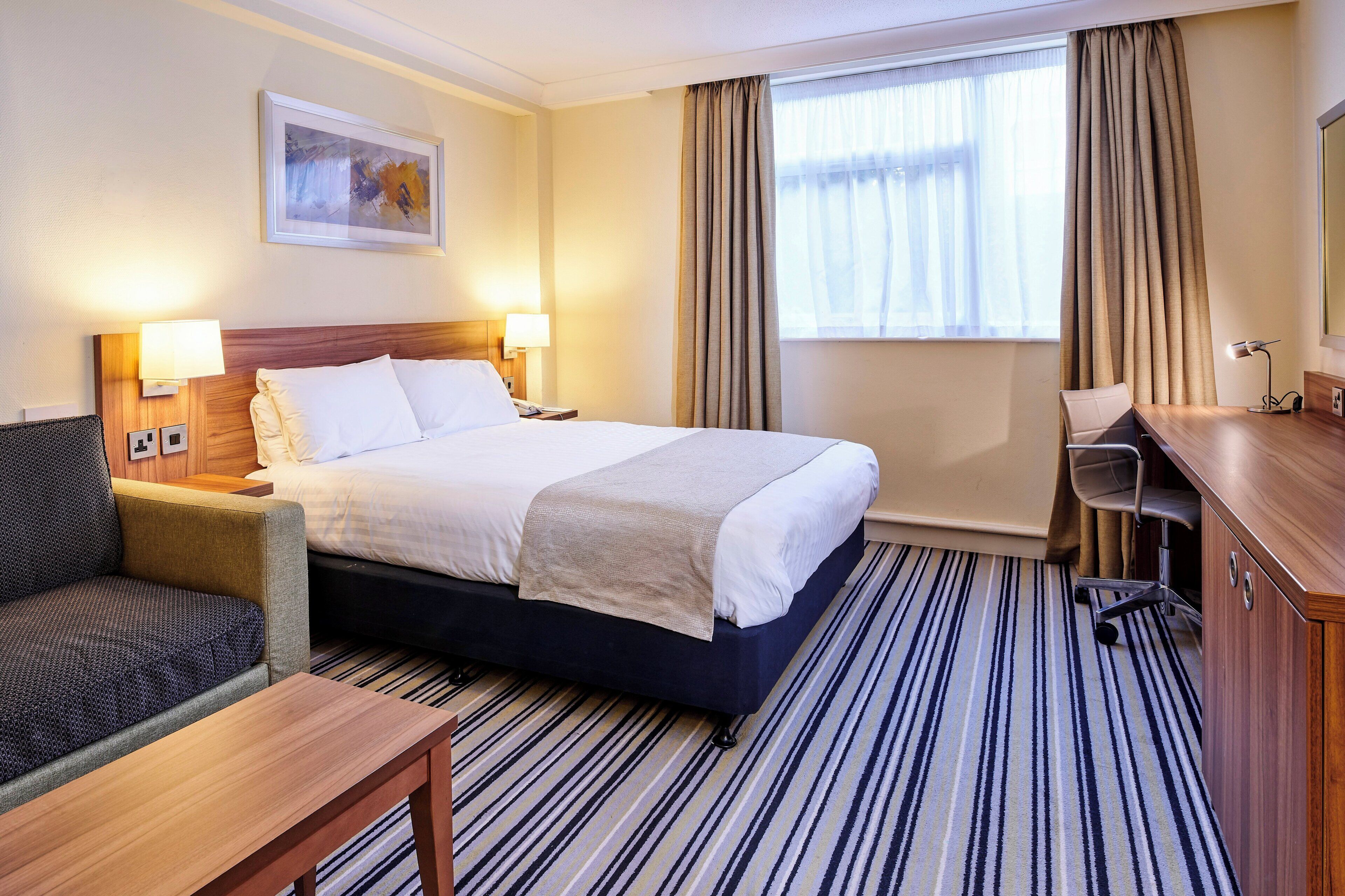 A hotel room with a bed, two bedside lamps, a desk, a chair, a sofa, a window with curtains, and a painting on the wall.