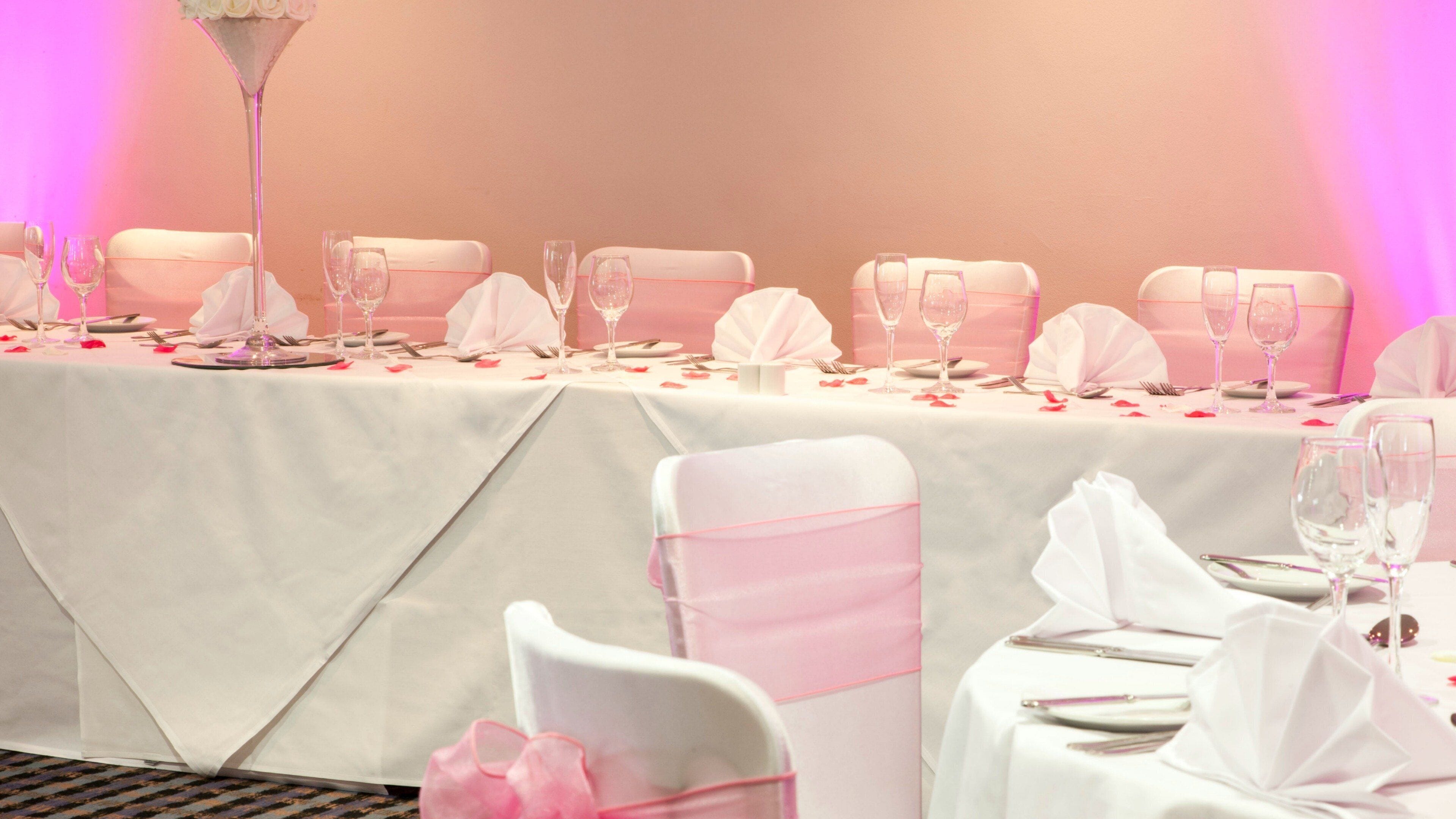 A banquet table set for a formal event with white tablecloths, pink sashes, and clear glassware.