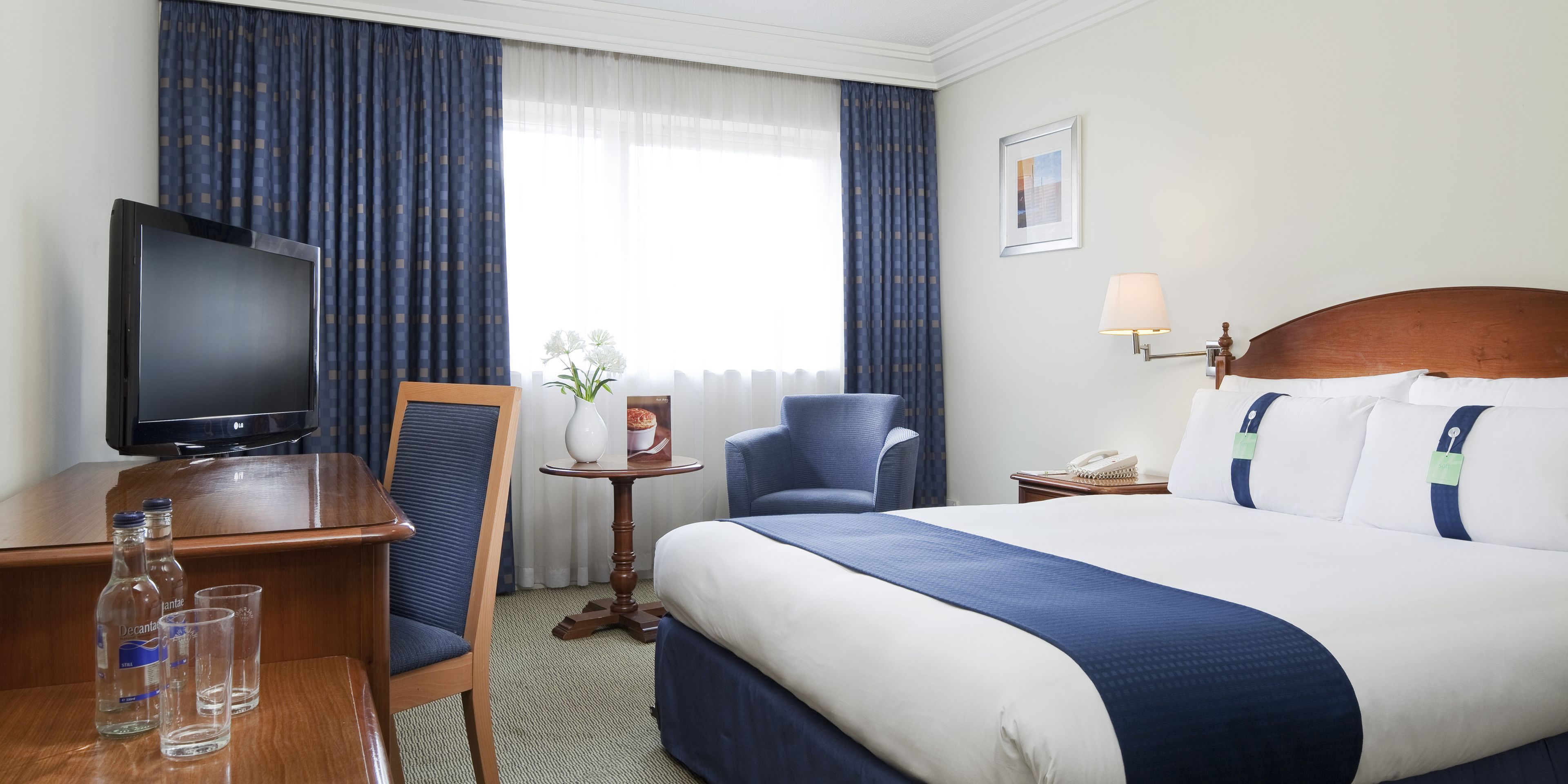 A hotel room with a large bed, a television, a desk with a chair, a small table with a vase and a picture frame, and a lamp.