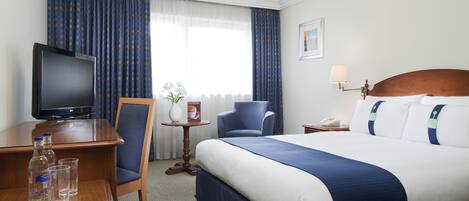 A hotel room with a large bed, a television, a desk with a chair, a small table with a vase and a picture frame, and a lamp.