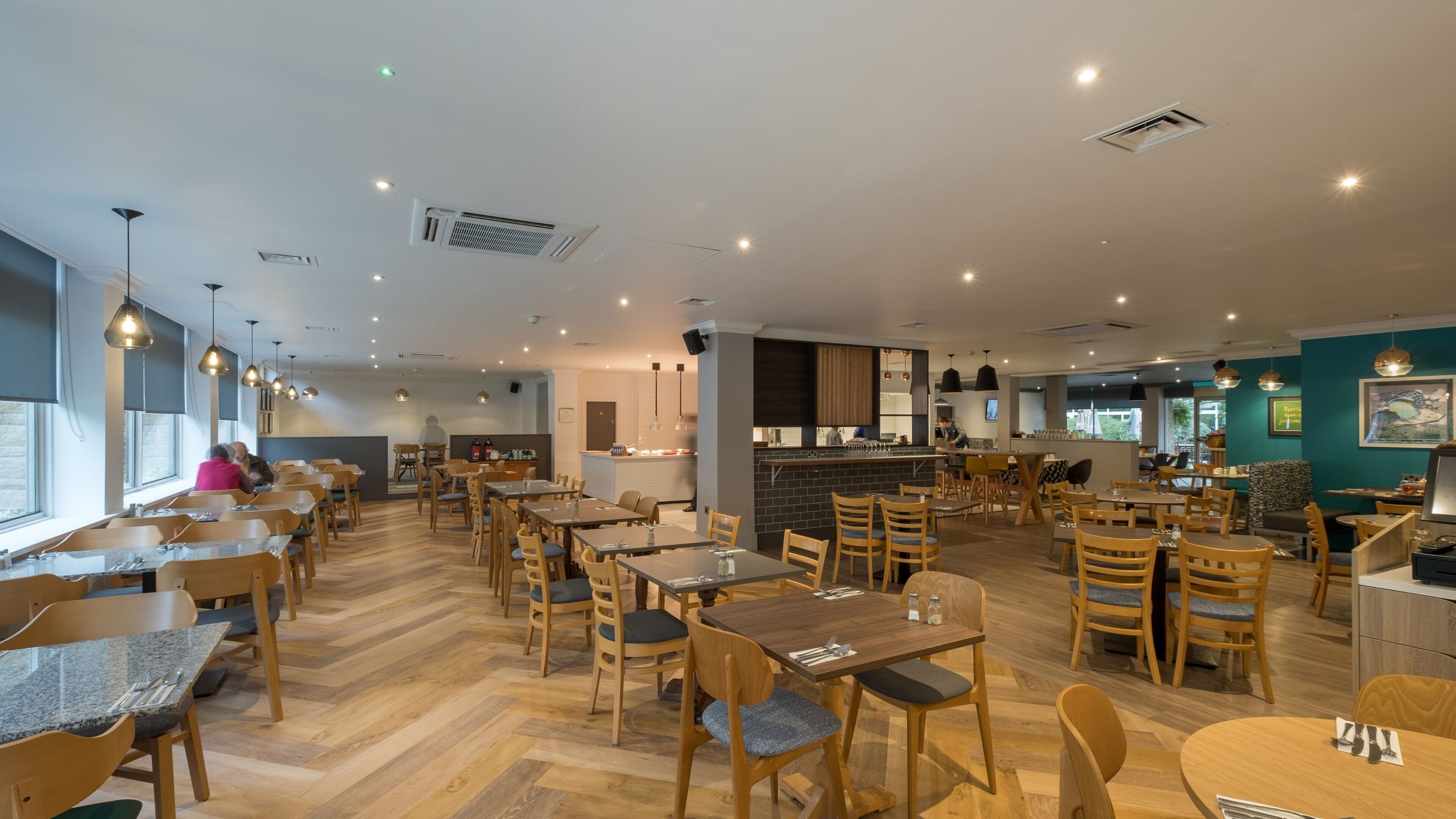 A spacious restaurant with multiple dining areas, wooden tables and chairs, and a kitchen visible in the background.