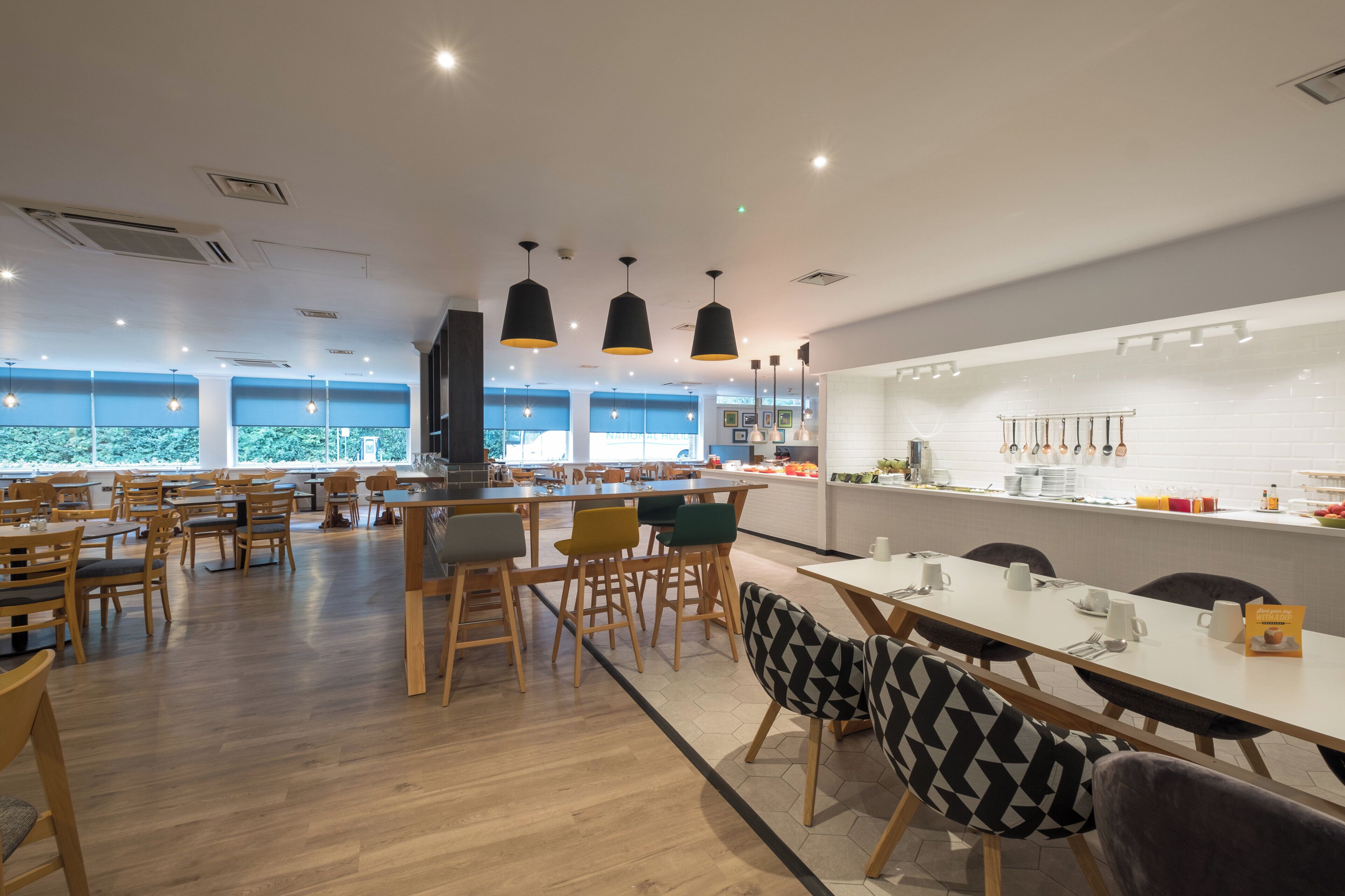 A modern dining area with wooden tables and chairs, pendant lighting, and a view of the outdoors through large windows.