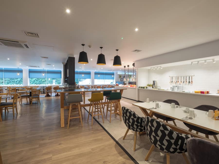A modern dining area with wooden tables and chairs, pendant lighting, and a view of the outdoors through large windows.