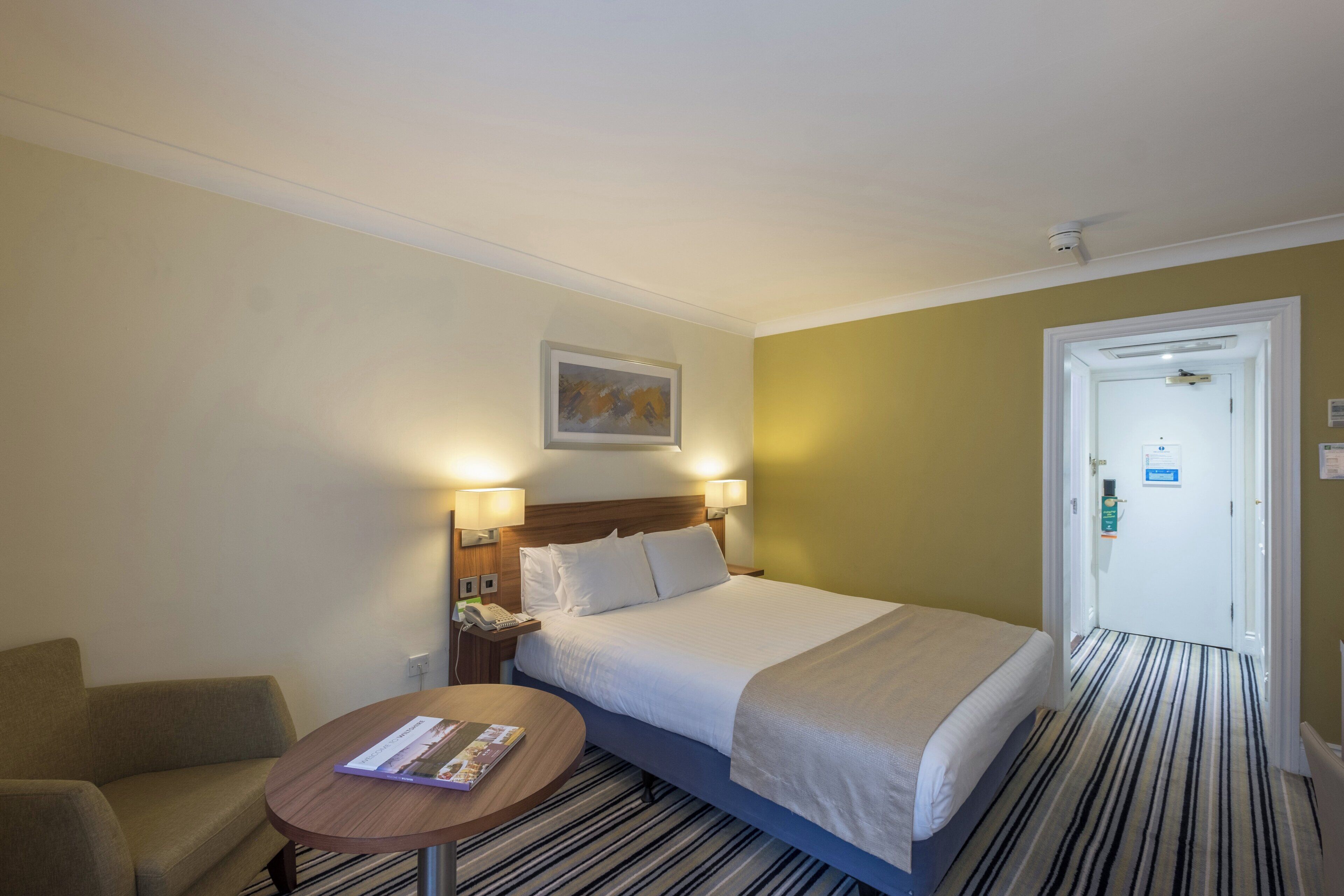 A hotel room with a bed, bedside lamps, a small table with a magazine, a painting on the wall, and a door with a sign.